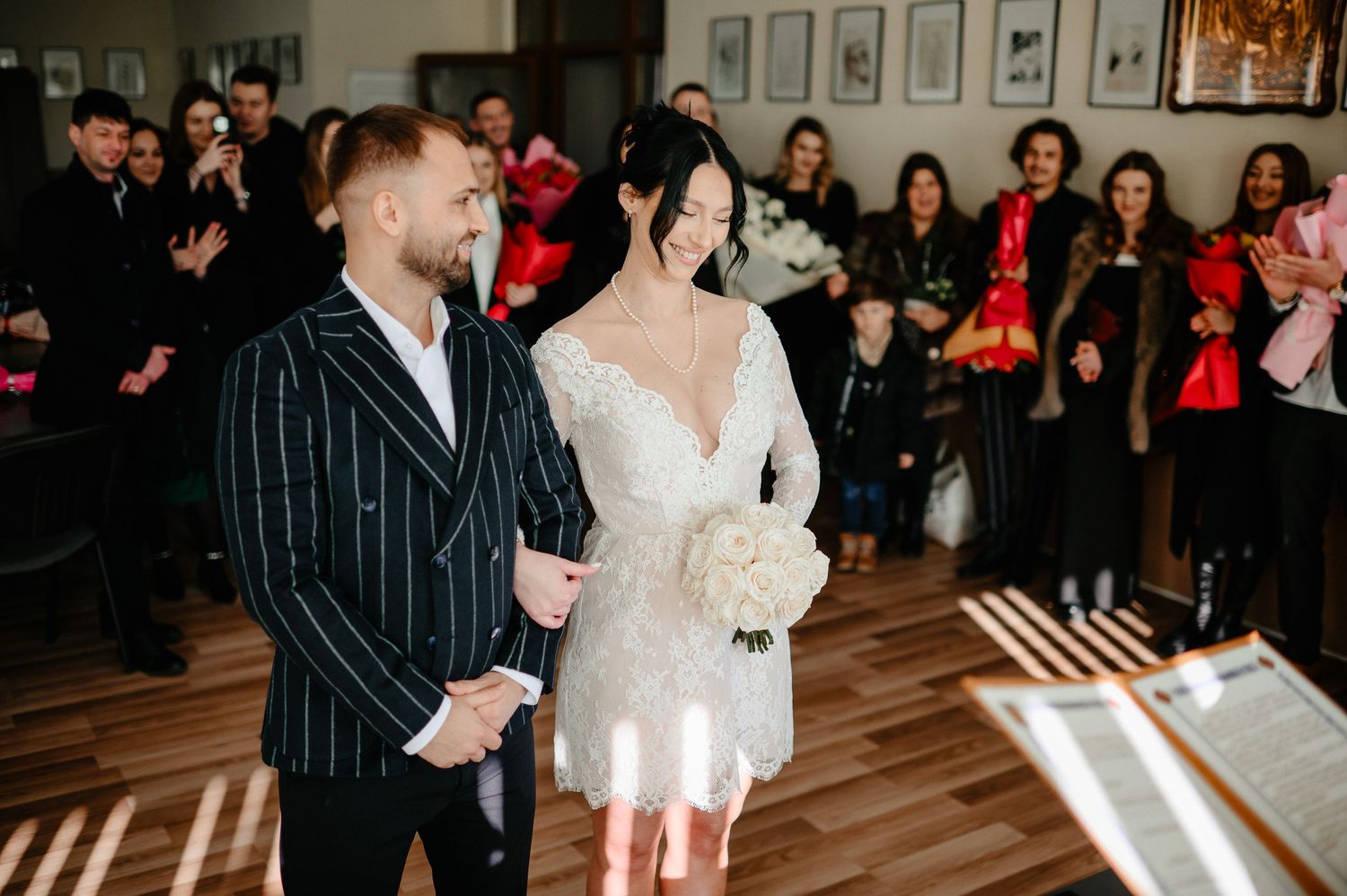 Bride in short lace dress and groom in pinstripe suit exchange vows at intimate wedding ceremony.