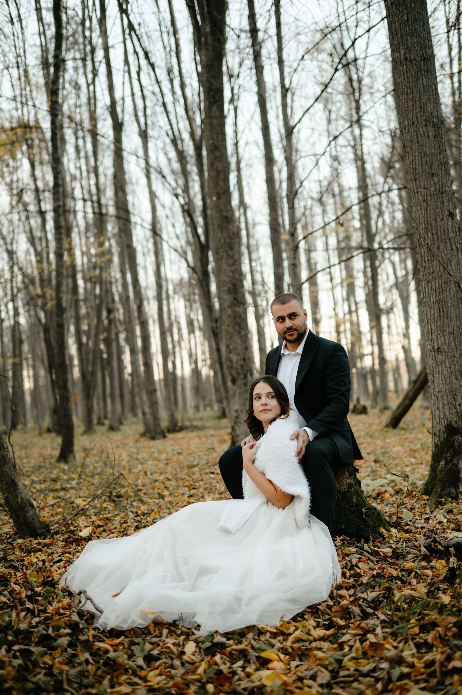 Bride in white gown sits on autumn leaves leaning against groom in black suit in a bare forest setting.