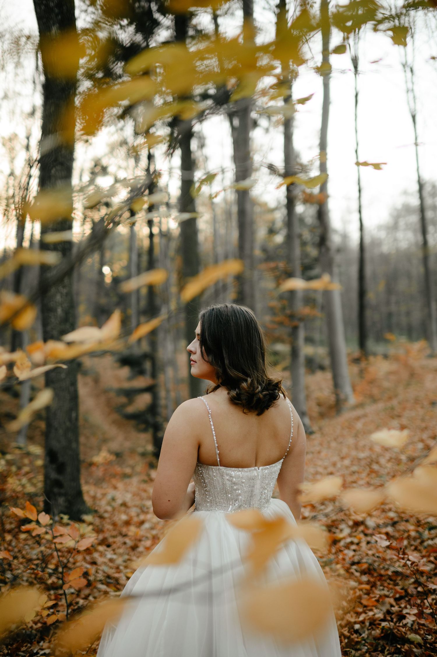 Bride in white gown standing in autumn forest with falling yellow leaves.