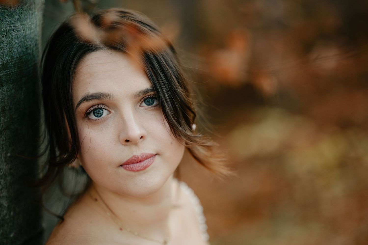 Young woman with green eyes and brown hair poses against a tree in an autumn forest setting.