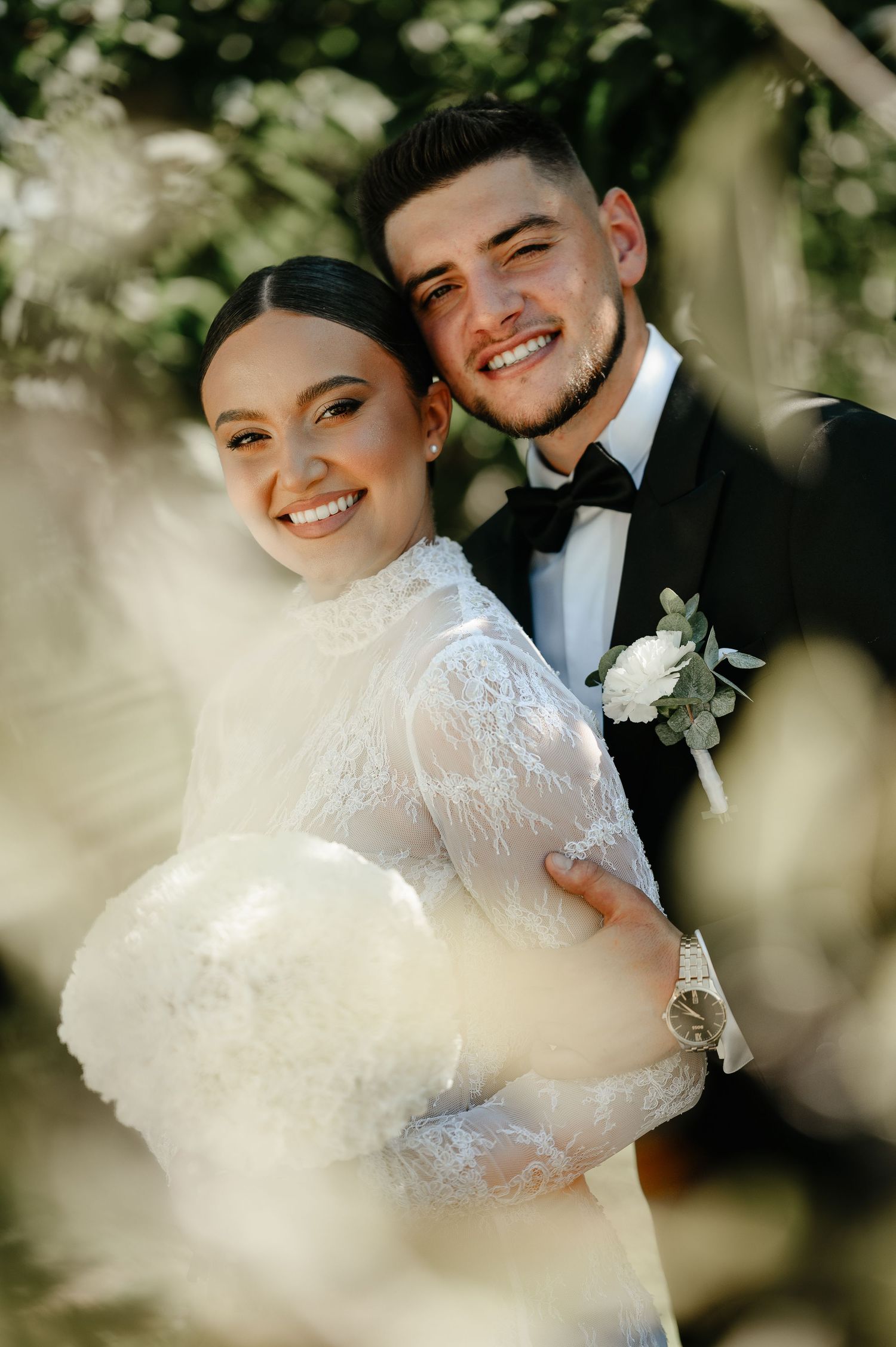 A smiling bride in lace gown holds white bouquet as groom in black tuxedo embraces her outdoors.