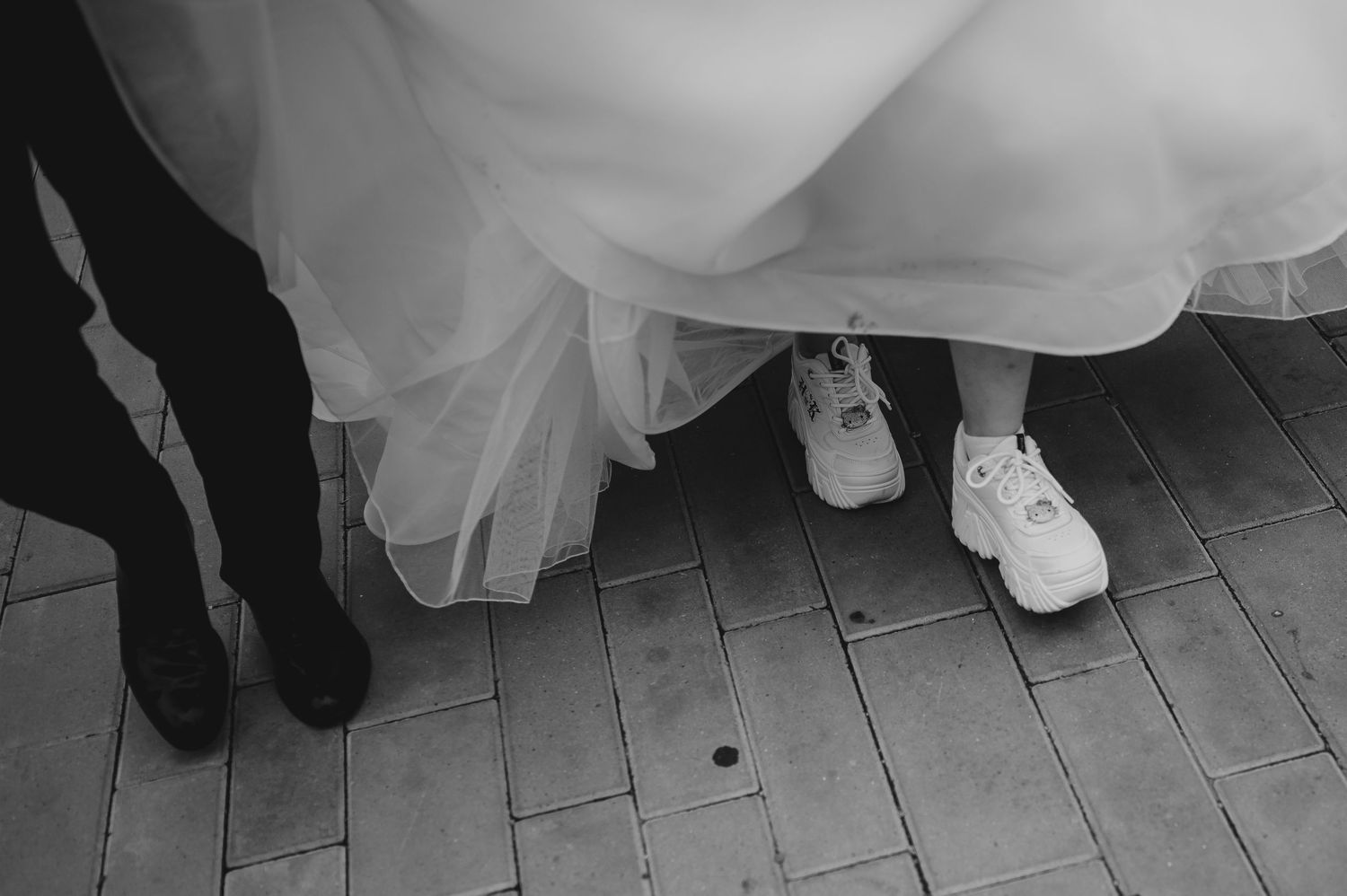 Black and white photo of bride in wedding dress wearing white sneakers beside groom in black boots.