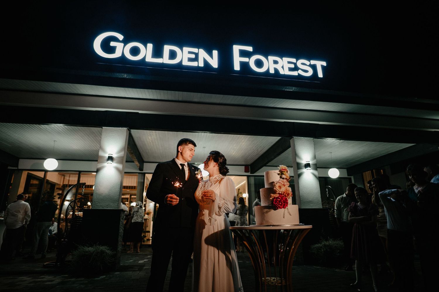 Bride and groom cutting wedding cake outside Golden Forest venue at night.