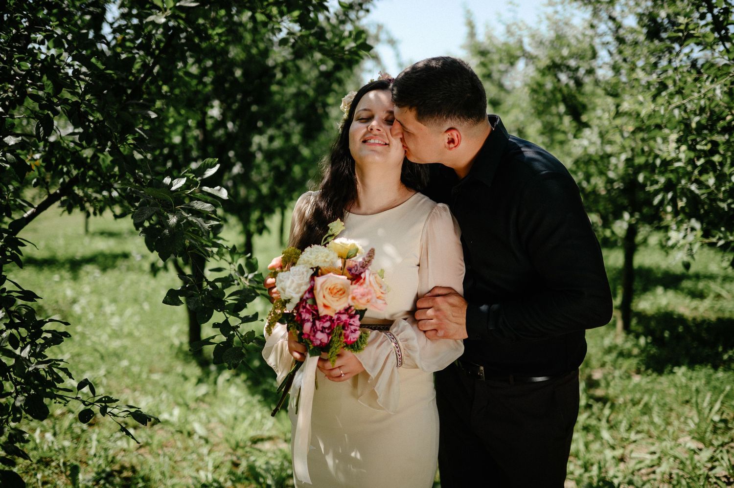 Couple embracing in an orchard, bride holding colorful bouquet, groom kissing her cheek on their wedding day.