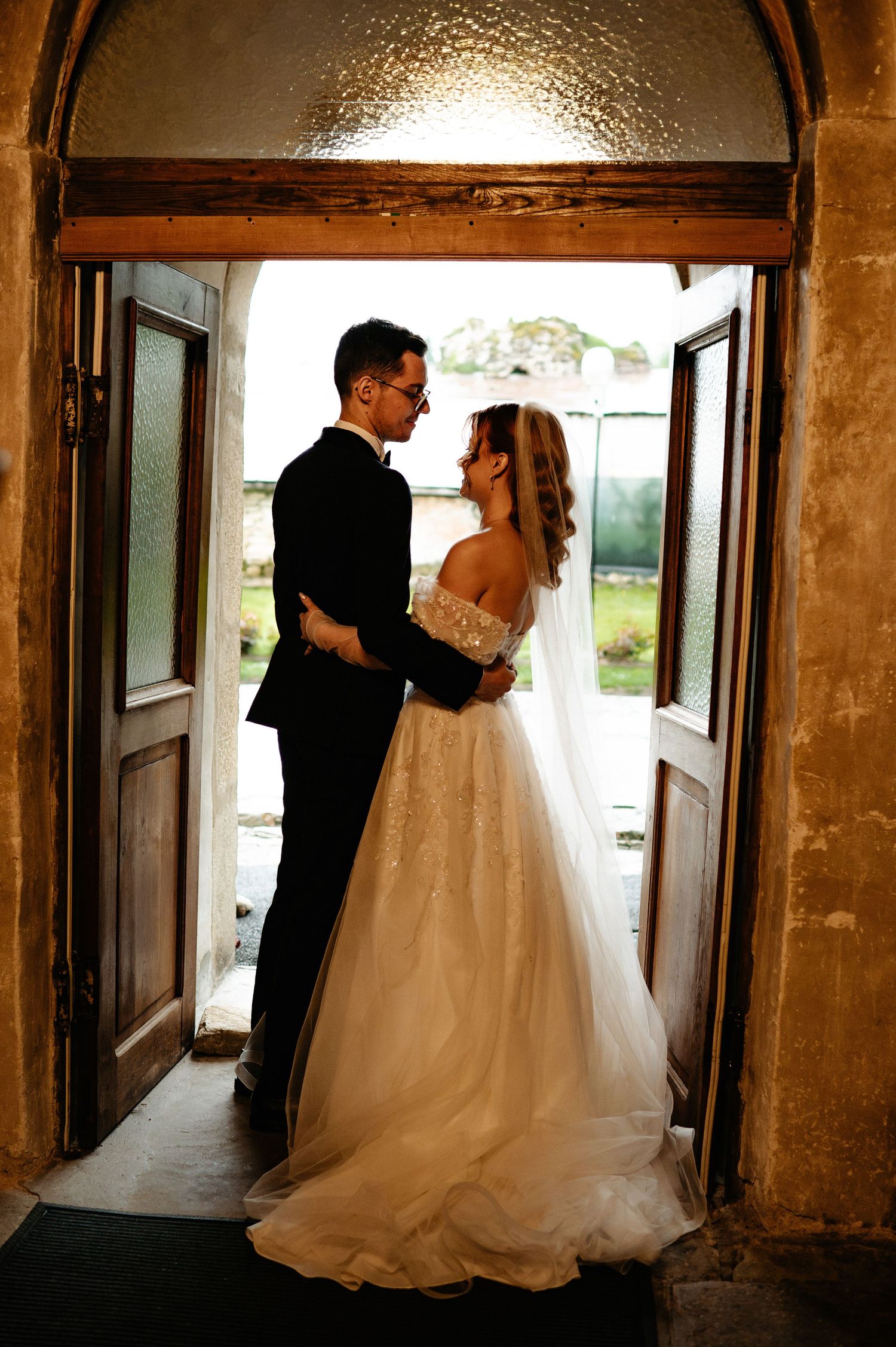 Bride and groom share a romantic moment in a rustic doorway, bride in lace gown with veil, groom in black suit.