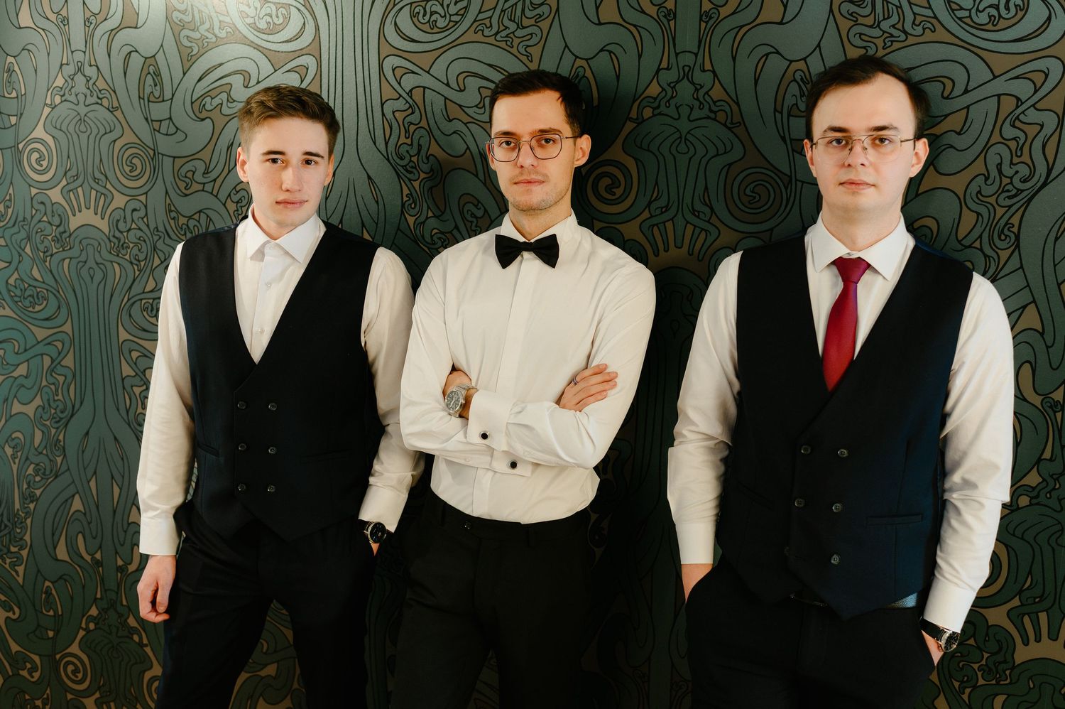 Three young men in formal black vests and white shirts pose together against a decorative wall.