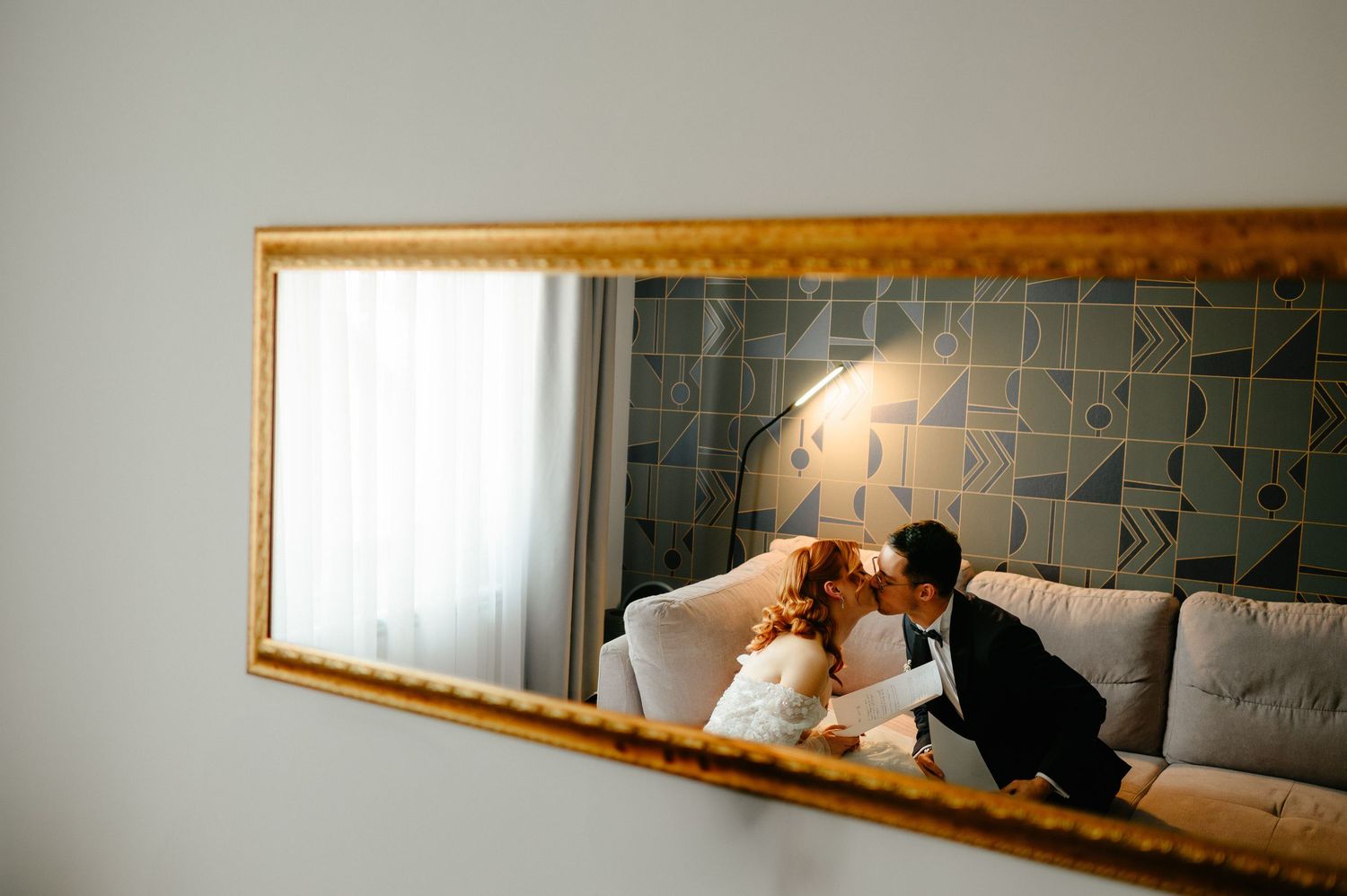 Bride and groom sharing a romantic kiss reflected in a gold-framed mirror on a elegant sofa.