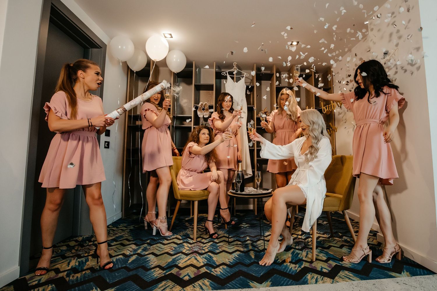 Bridesmaids in pink dresses celebrate with confetti and champagne around bride in white during wedding prep.