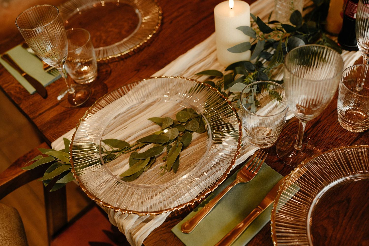 Elegant table setting with ribbed glass charger plates, crystal glassware, candles, and fresh greenery centerpiece.