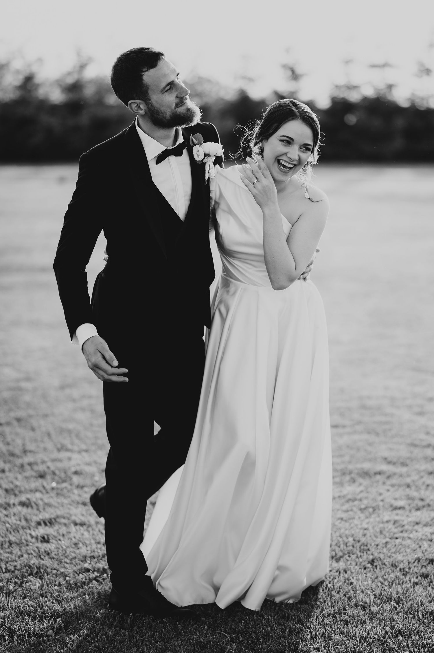 Laughing bride in flowing white gown leans on smiling groom in black tuxedo on grassy field at dusk.