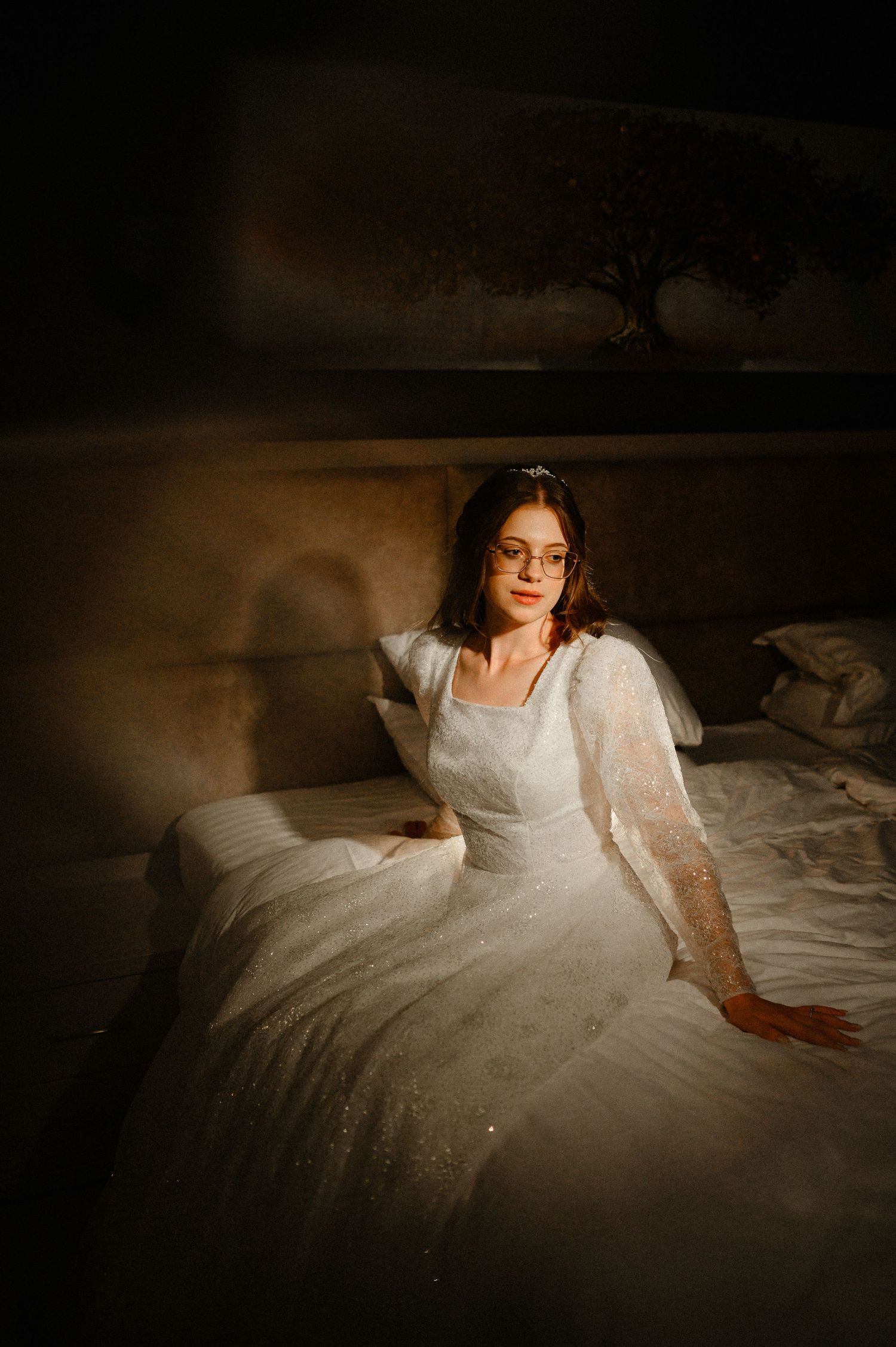Bride in white sparkle wedding dress and glasses sitting on bed in warm golden light.