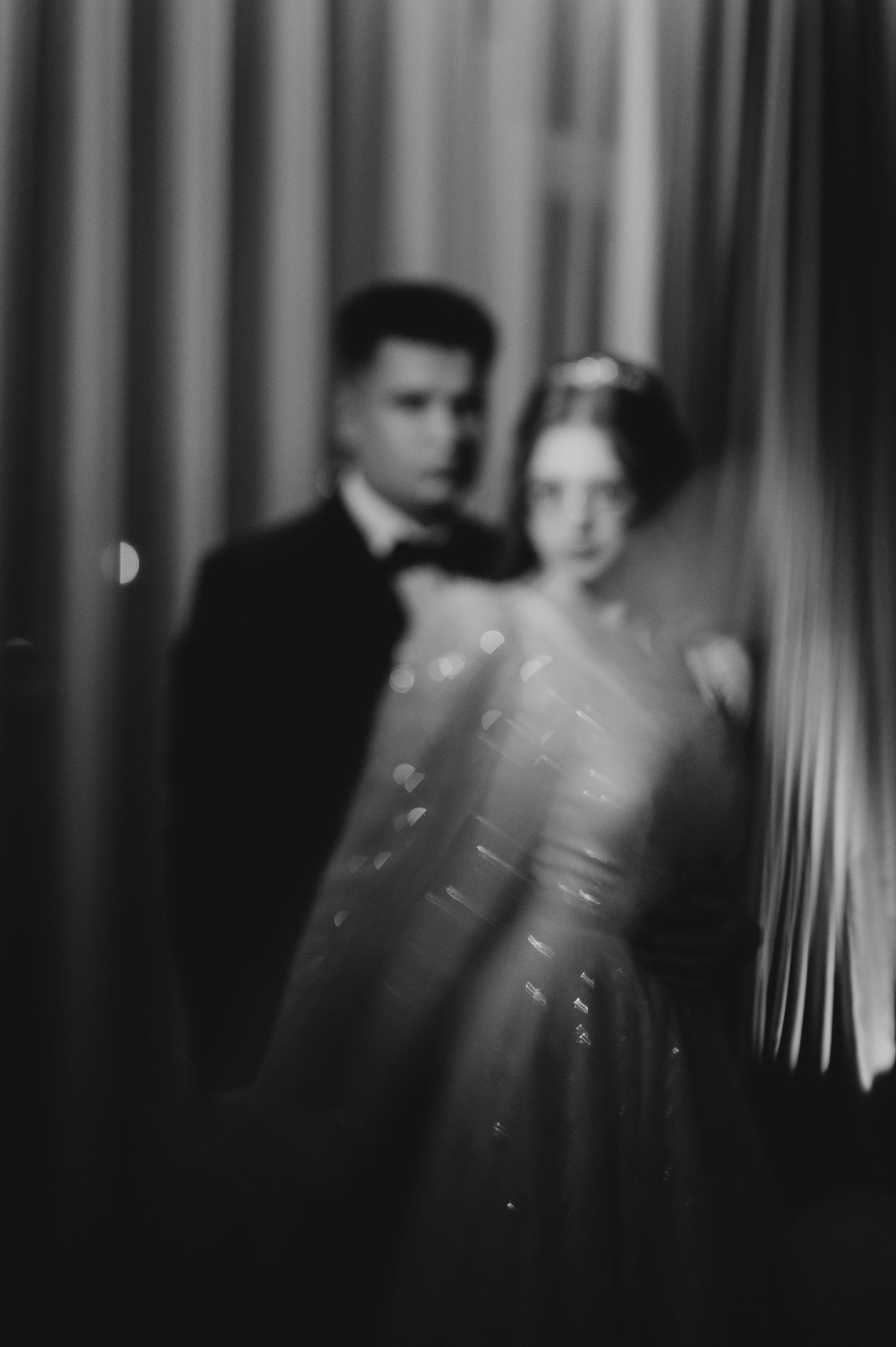 Elegant black and white blurred photo of a bride and groom standing behind sheer curtains.