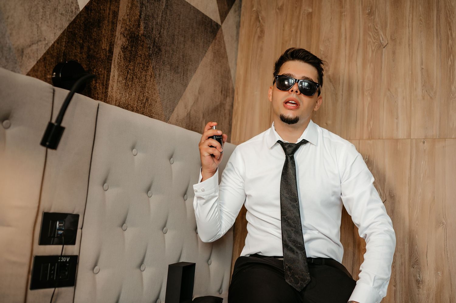 Stylish man in white shirt and tie wearing sunglasses holding a cigar in a modern interior.