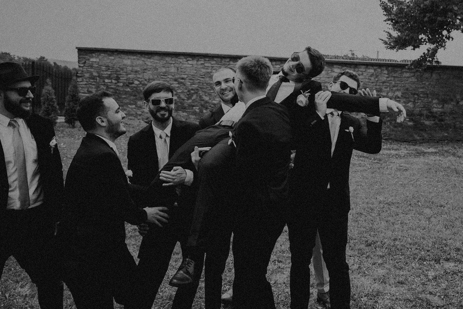 Groomsmen in black suits laughing and celebrating together in a candid black and white outdoor wedding photo.