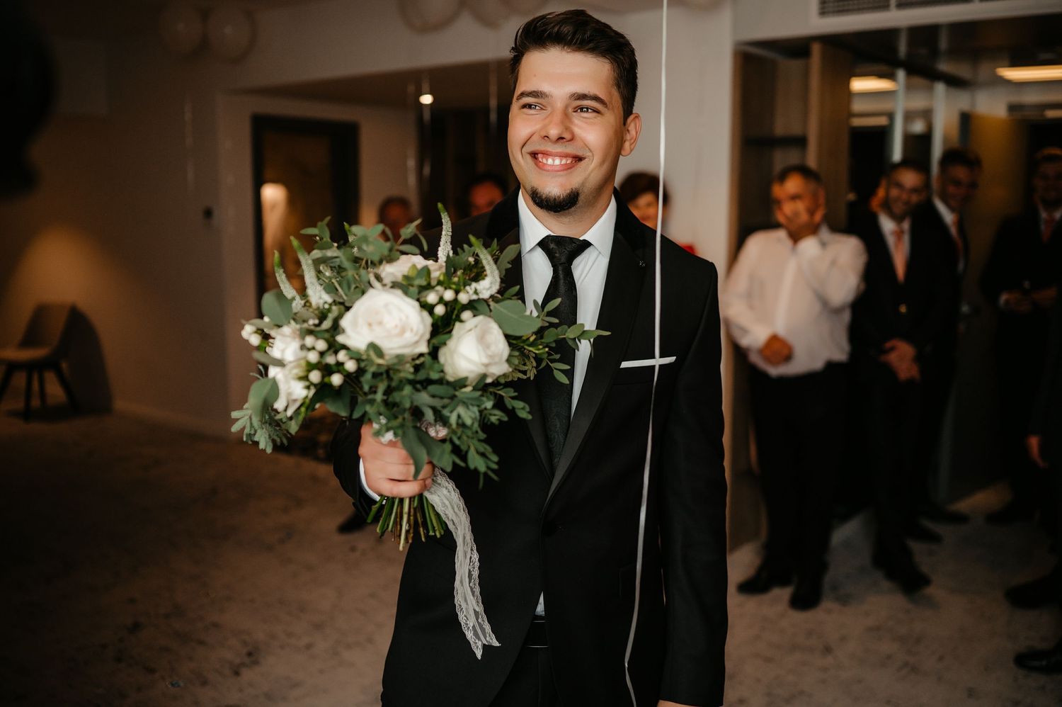 Smiling groom in black tuxedo holding white and green bridal bouquet at wedding ceremony.