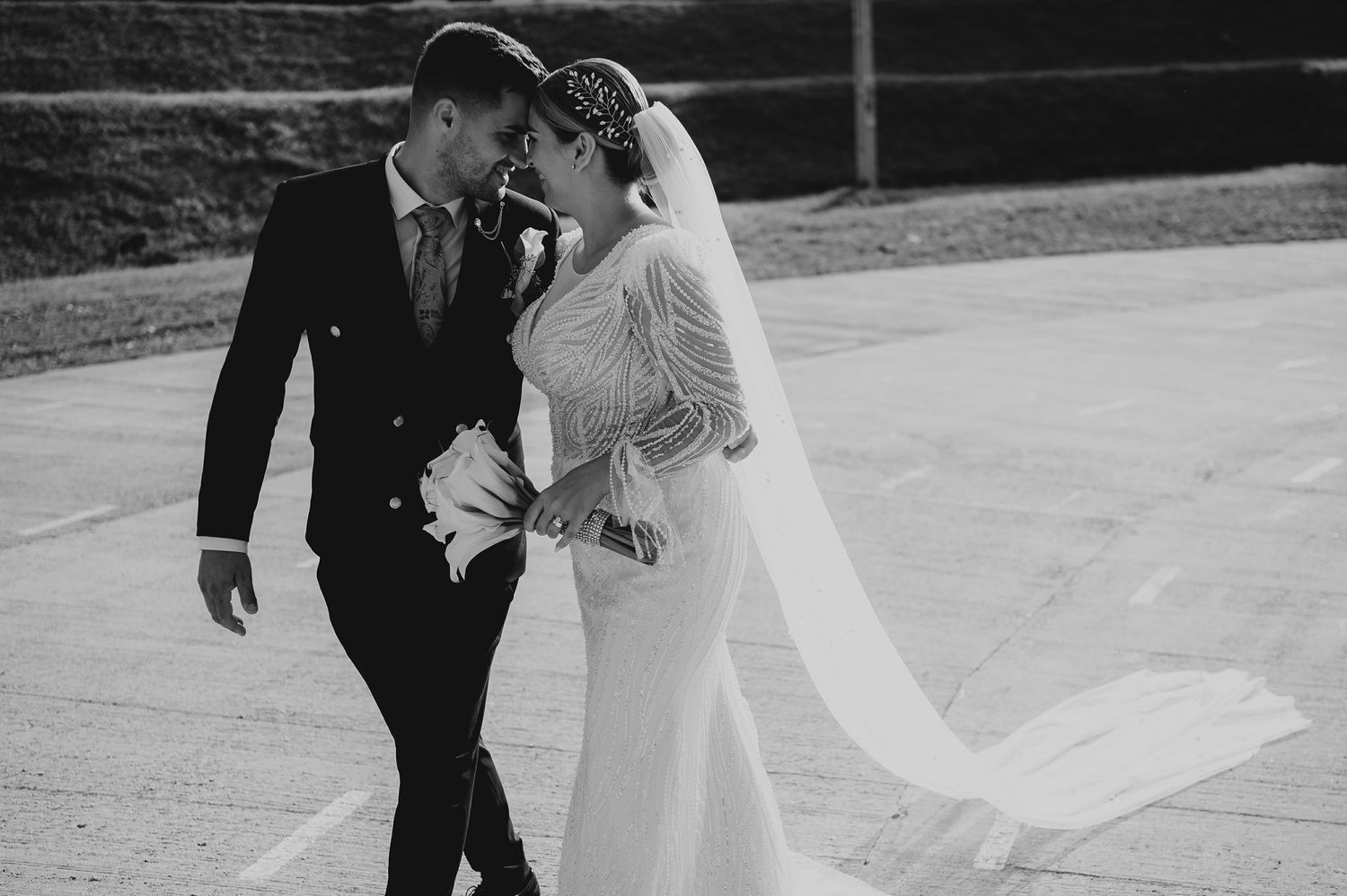 Black and white photo of bride and groom walking together outdoors, bride's veil flowing in the breeze.