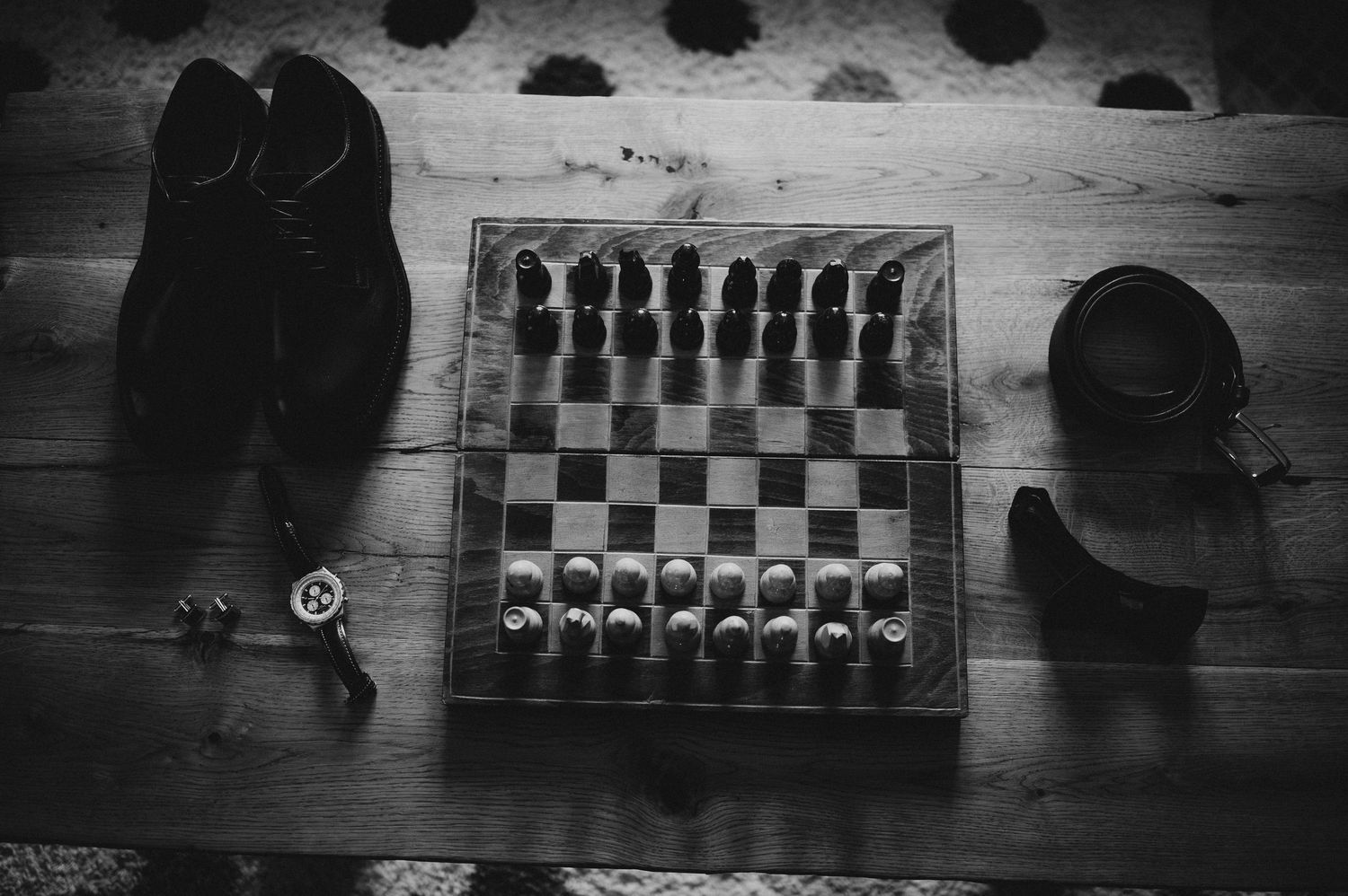 Black and white overhead view of a chess board set up for play on a wooden floor beside dress shoes.