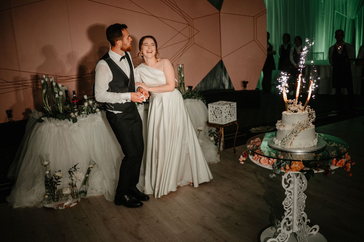 Happy couple in wedding attire smile together beside decorated white wedding cake with sparklers.