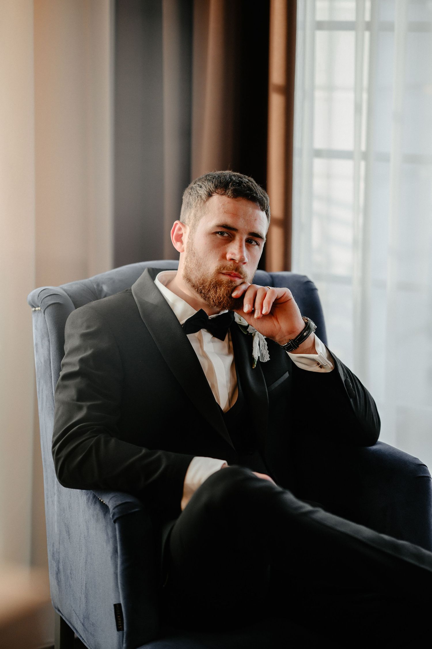 Handsome groom in black tuxedo with bow tie sits thoughtfully in velvet armchair by window.