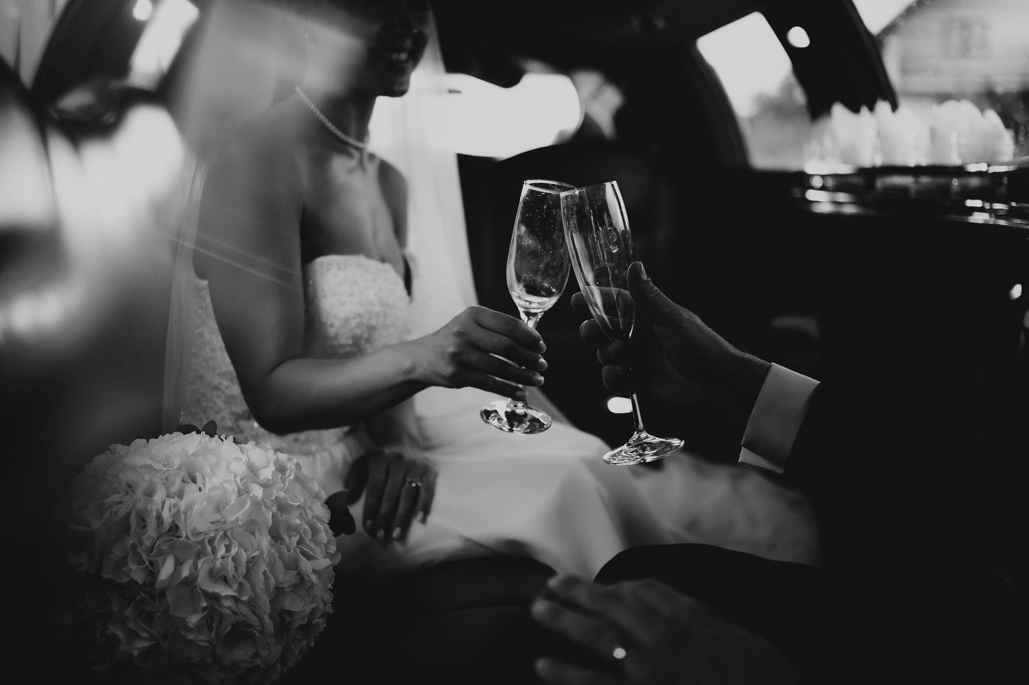 Elegant black and white close-up of bride and groom clinking champagne glasses in a limousine.