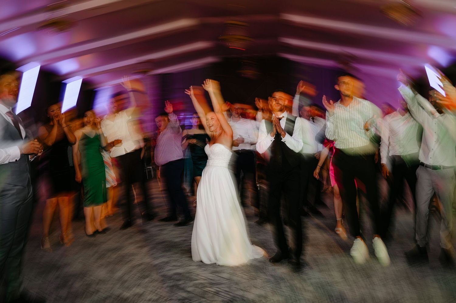 Motion blur photo of bride and groom dancing with raised arms at vibrant purple-lit wedding reception.