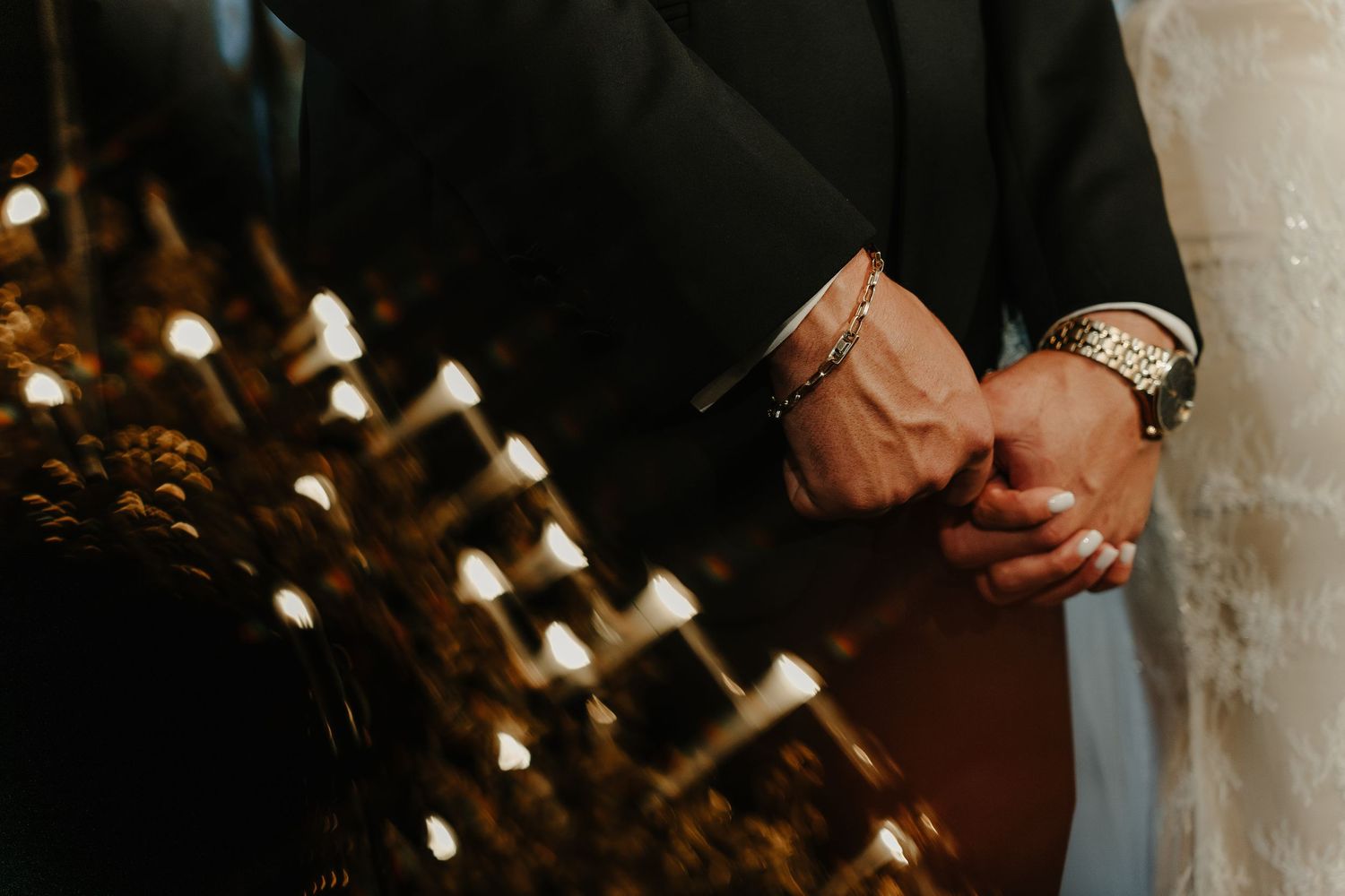 Close-up of groom and bride holding hands near golden candles during wedding ceremony.