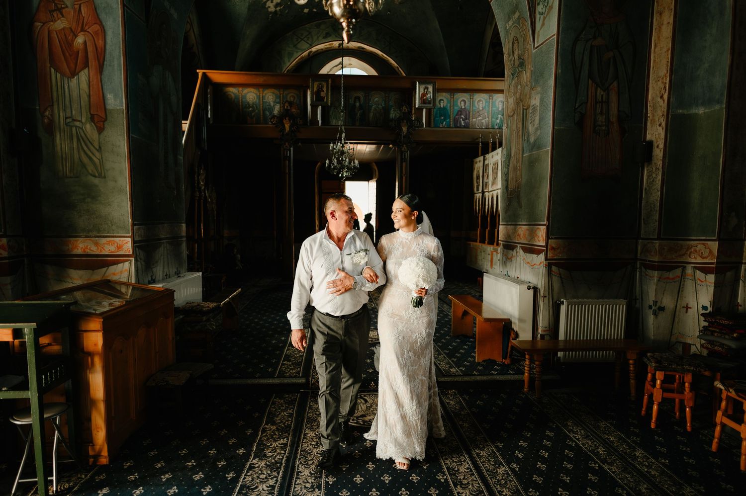 Bride in lace gown walking through ornate Orthodox church interior with escort.