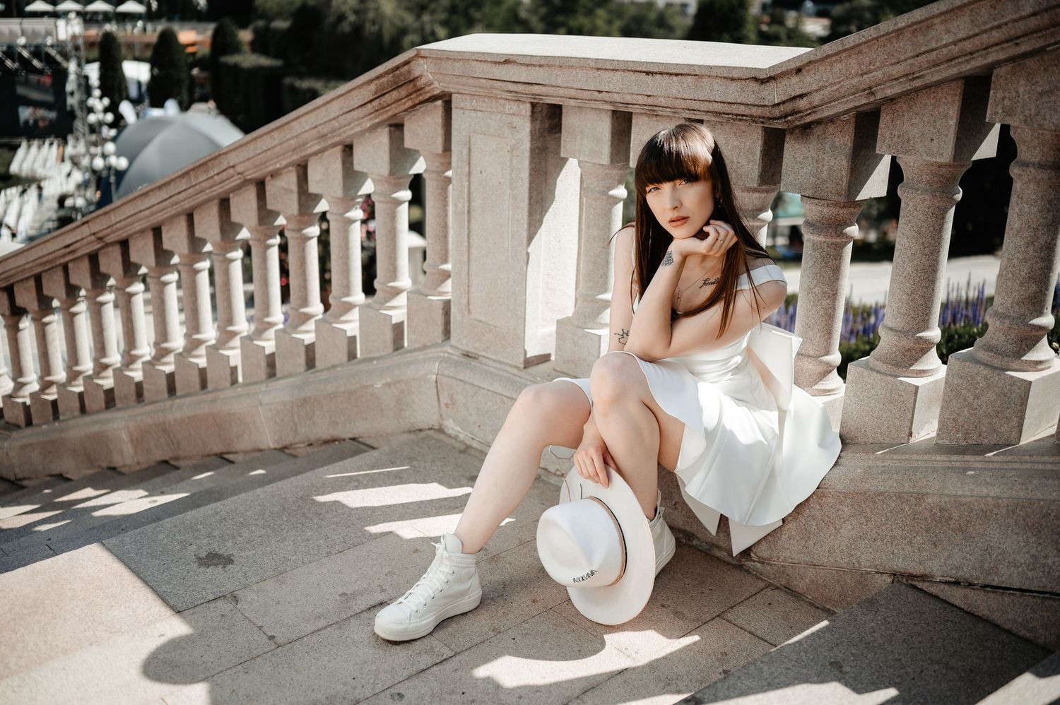 Young woman in white dress and socks sits on sunlit stone steps holding a white hat outdoors.