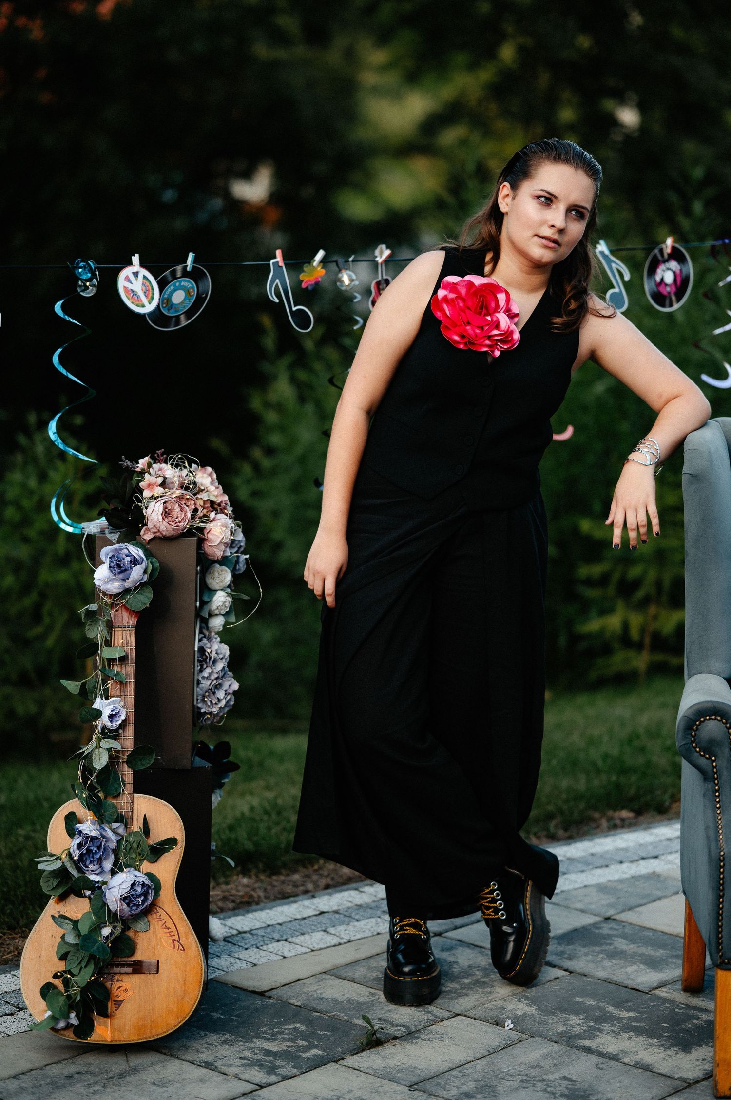 Young woman in black maxi dress with red corsage posing beside a floral-decorated guitar outdoors.