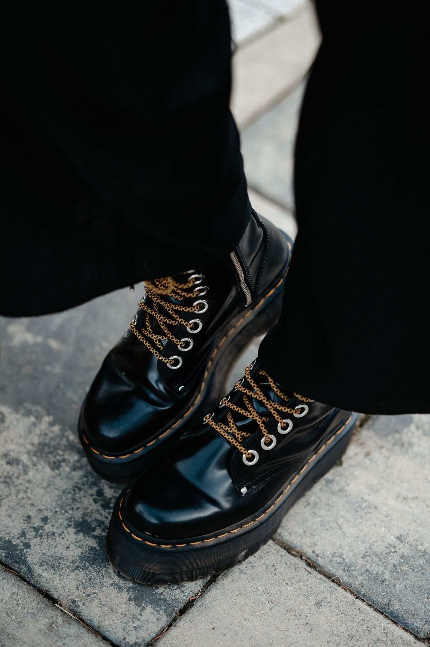 Close-up of black Dr. Martens platform boots with gold chain laces on cobblestone pavement.