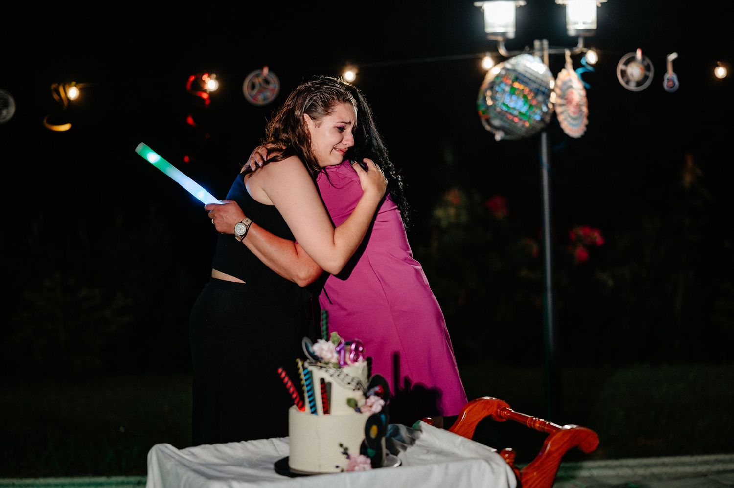 Two girls sharing a warm hug at a night birthday party with a decorated cake on the table nearby.