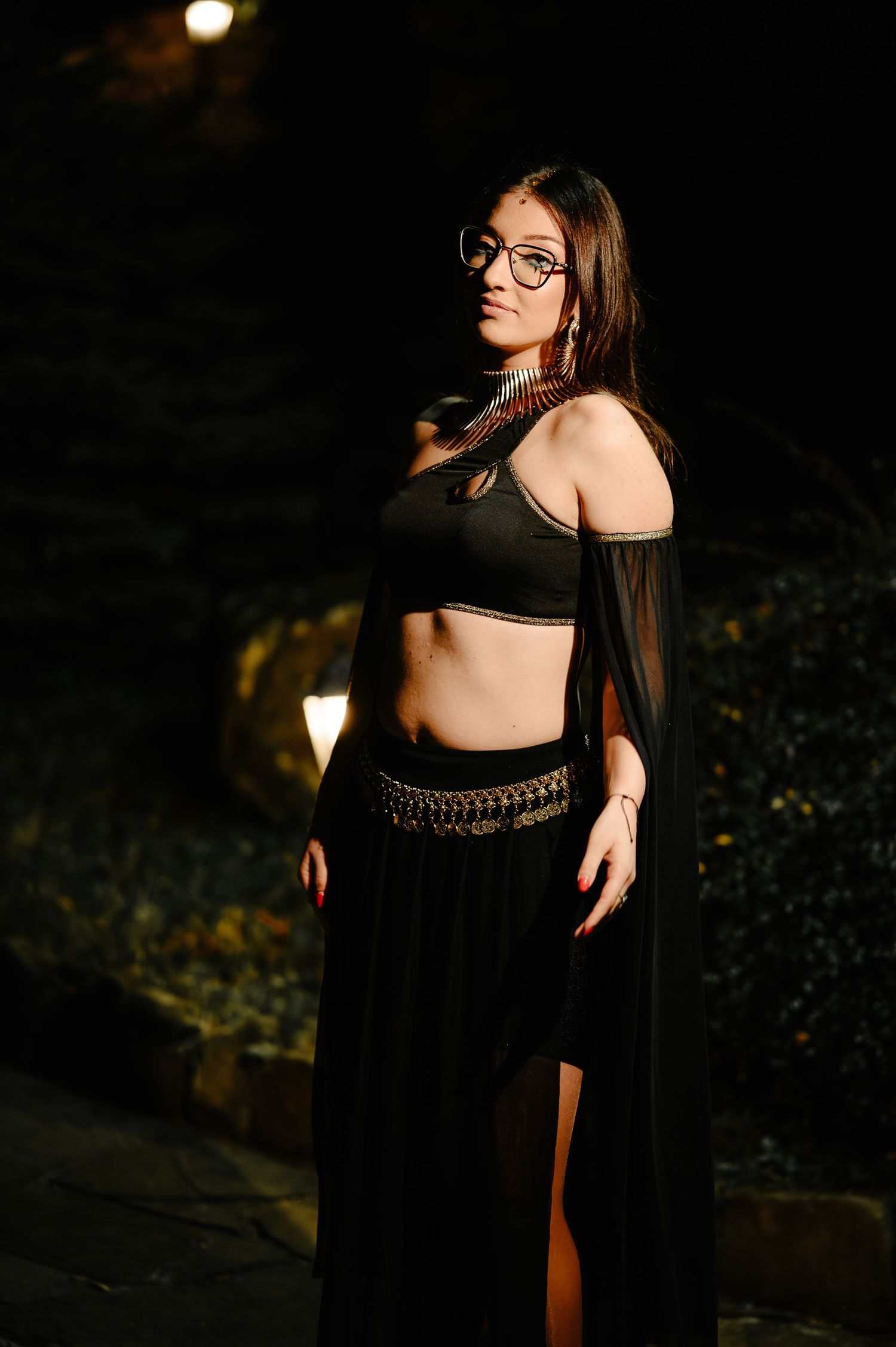 Woman in black crop top and maxi skirt with gold accessories poses outdoors at night.