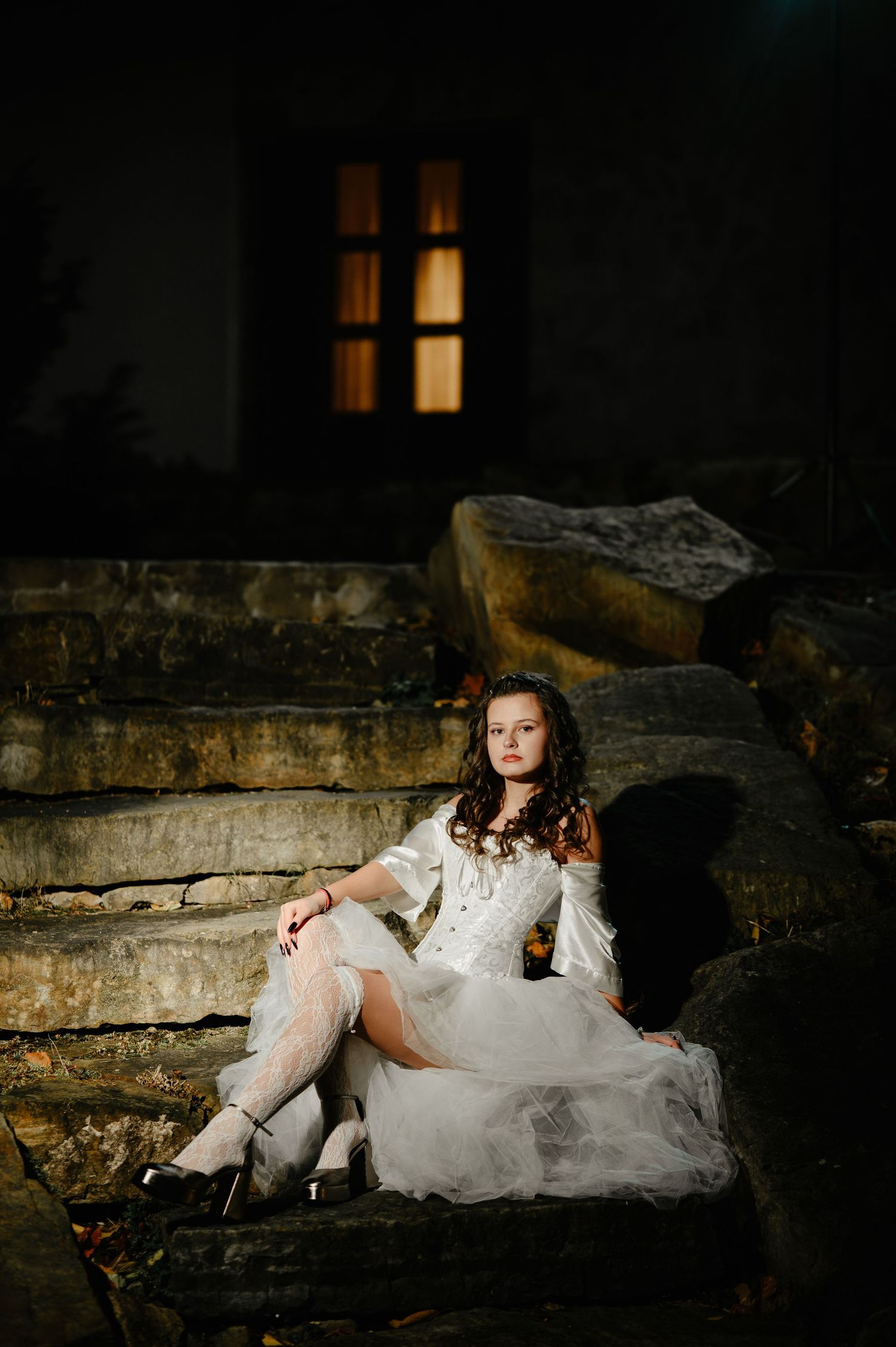 Young woman in white Halloween bride costume sitting on stone steps outside illuminated building at night.