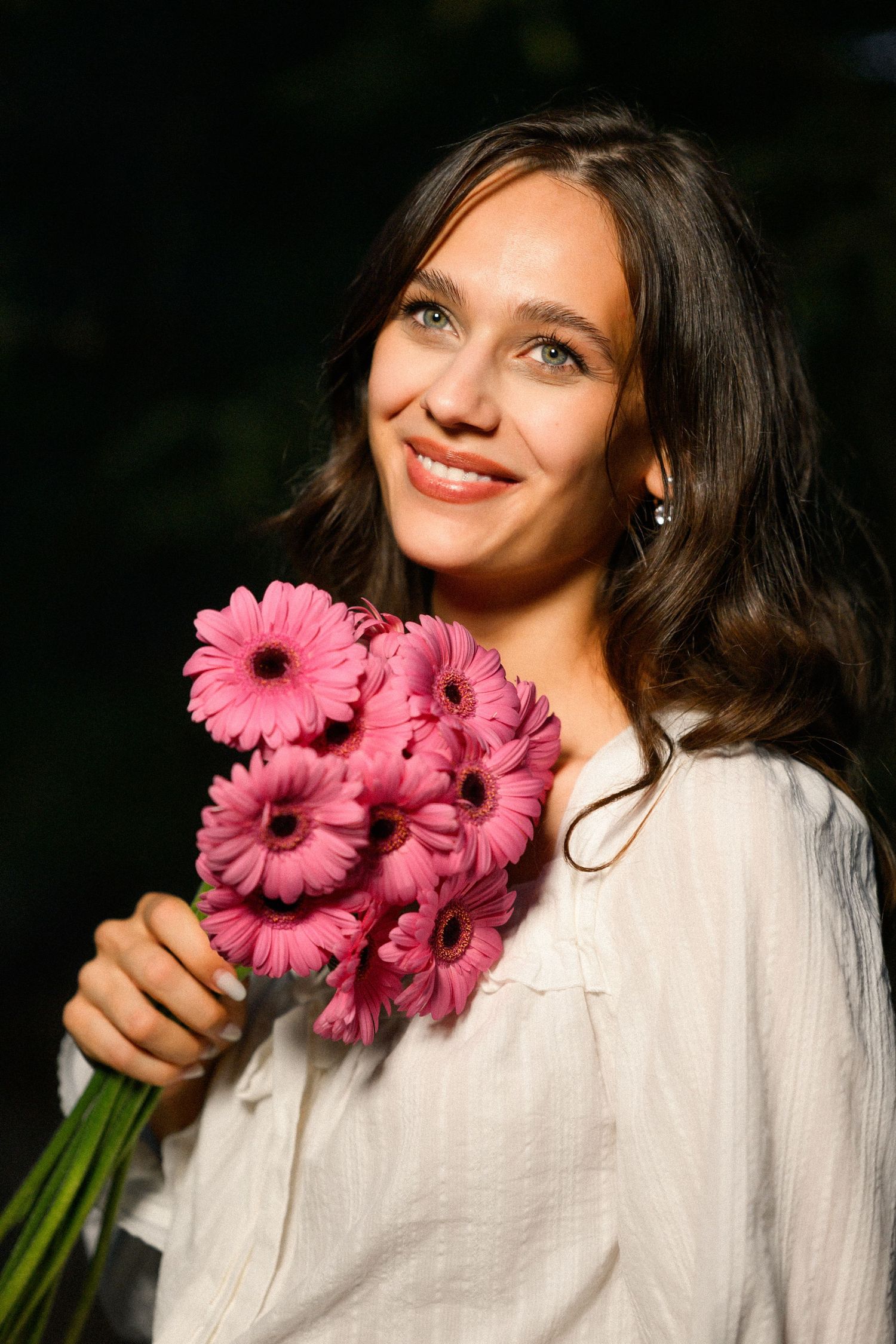 Smiling young woman with dark wavy hair holding a bouquet of pink gerbera daisies outdoors.