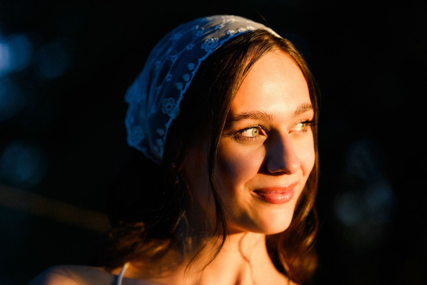 Young woman wearing a blue floral bandana smiles warmly in golden hour light against a dark background.