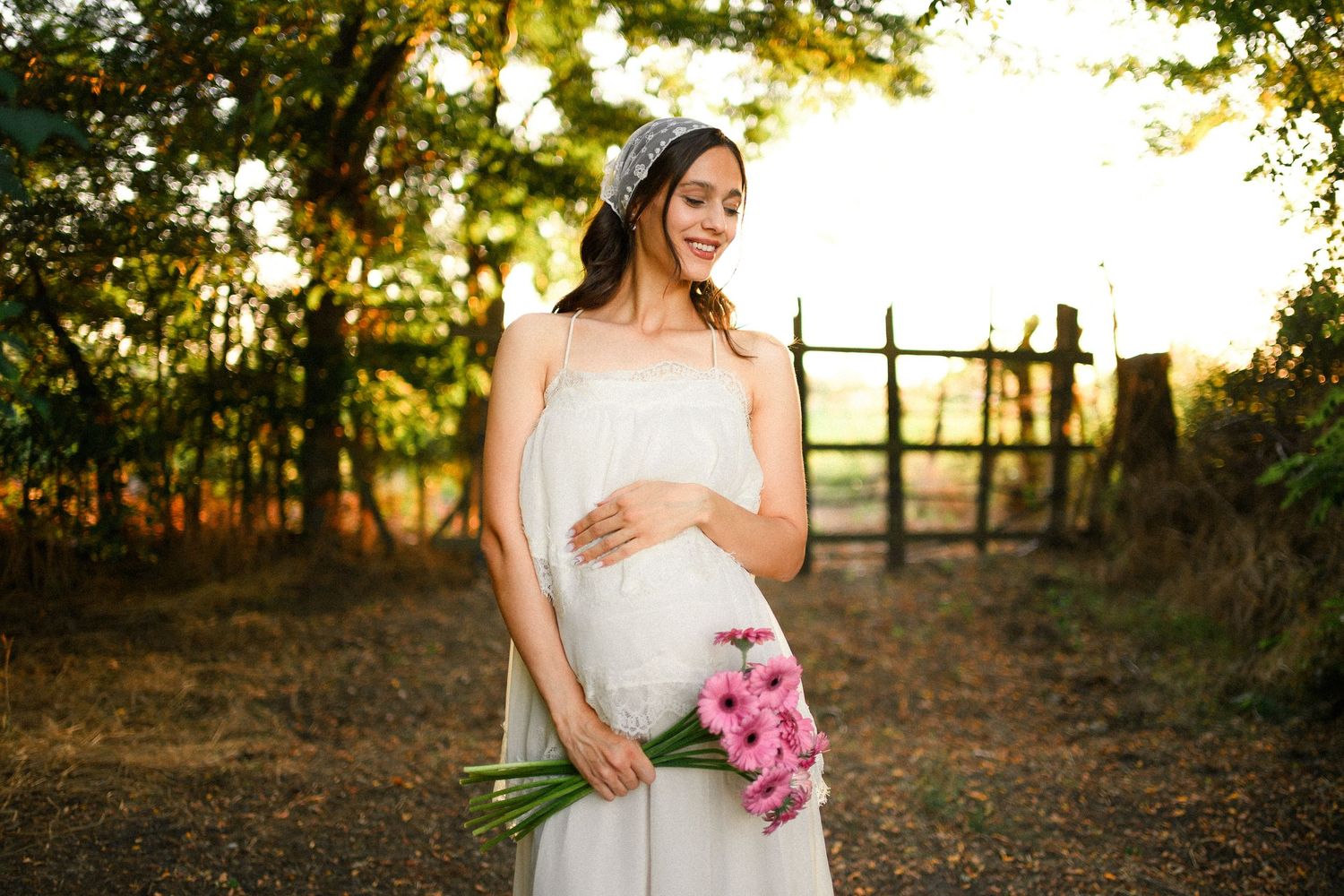 Pregnant woman in white dress holding pink flowers outdoors at golden hour, smiling serenely in a rustic garden setting.