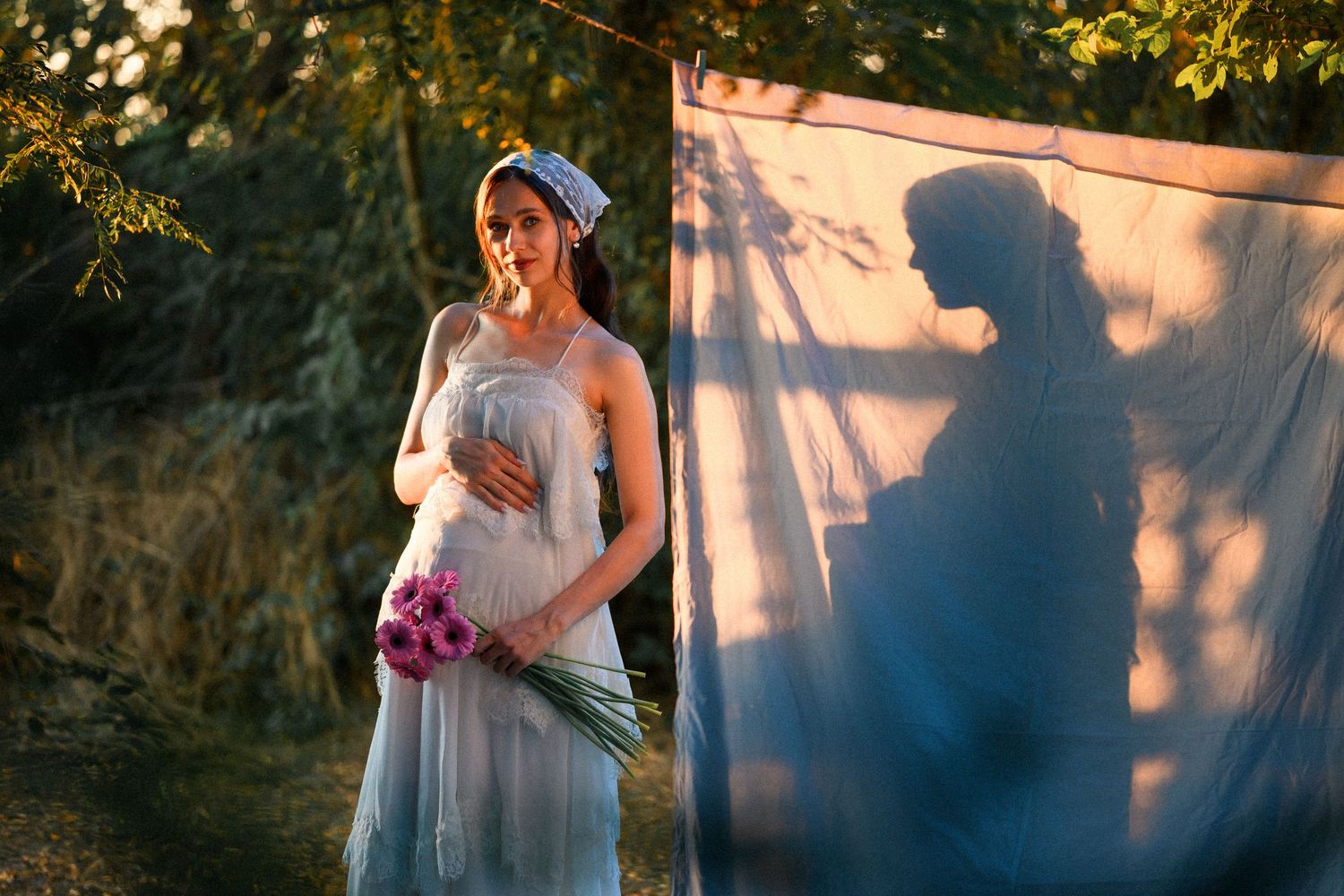 Pregnant woman in white dress holding pink flowers outdoors, with her shadow cast on a blue fabric backdrop.