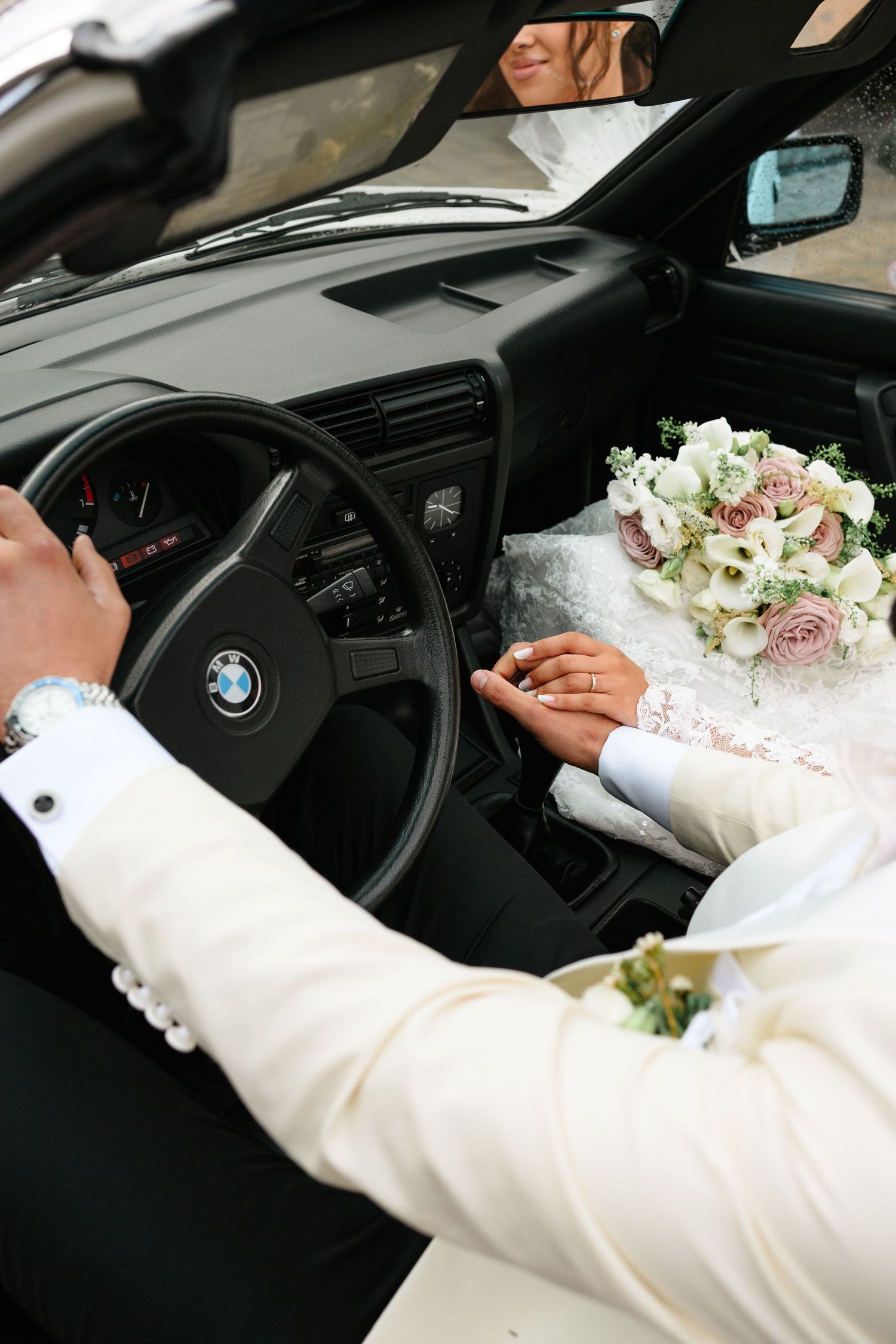 Newlyweds hold hands in a BMW car, with a bridal bouquet of pink and white roses on the dashboard.