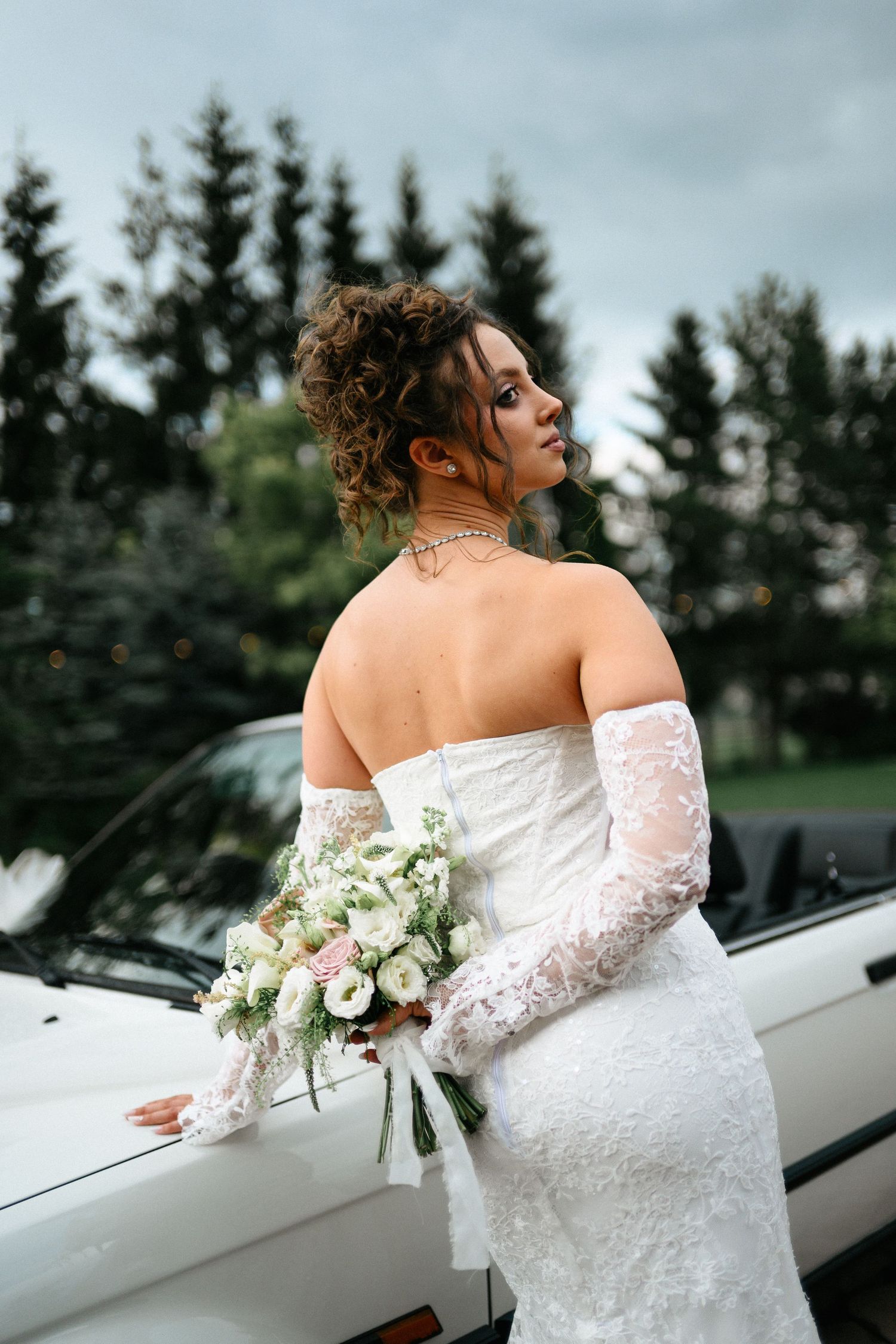 Bride in white lace off-shoulder gown holds bouquet, leaning on white car with evergreen trees behind.
