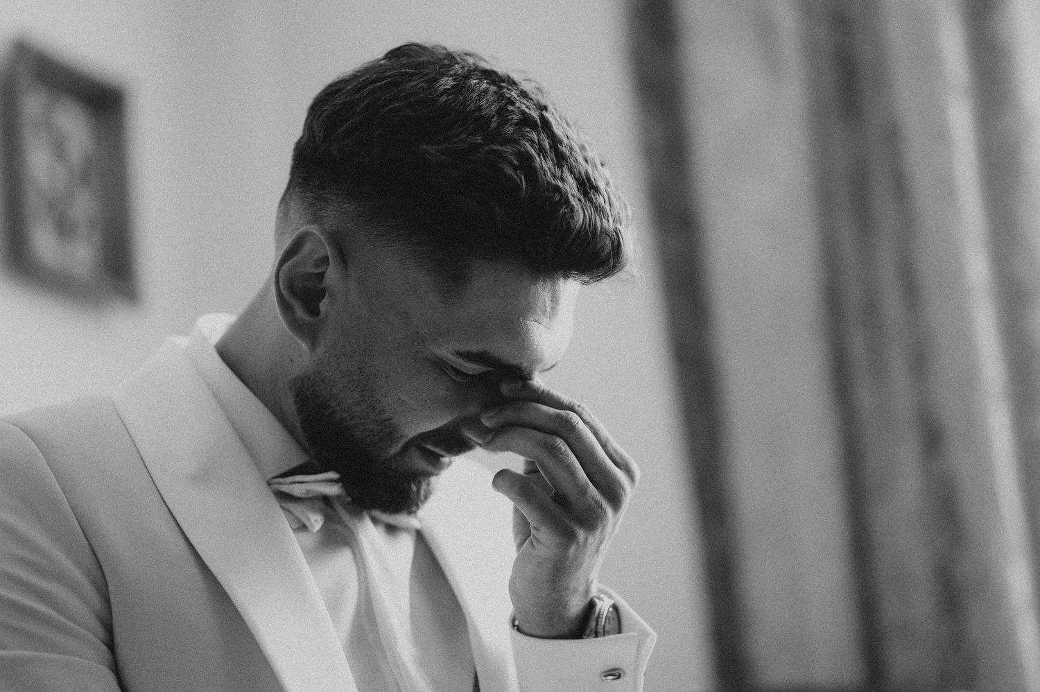 Emotional groom in a white tuxedo and bow tie wiping tears during wedding preparations, black and white photo.