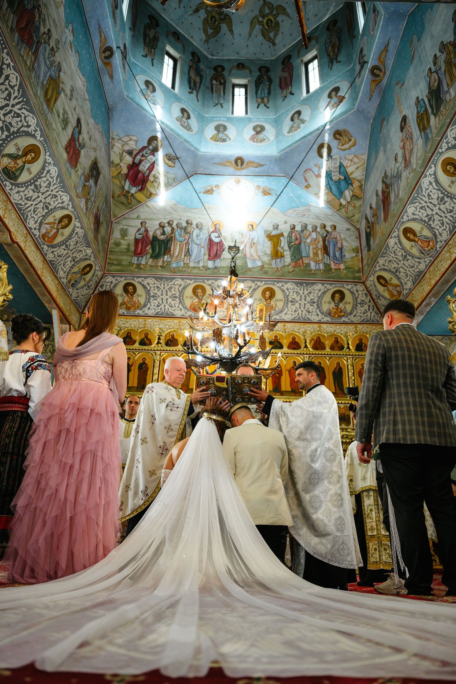 Orthodox wedding ceremony inside ornate Byzantine church with frescoed dome, bride in white gown with long train at altar.