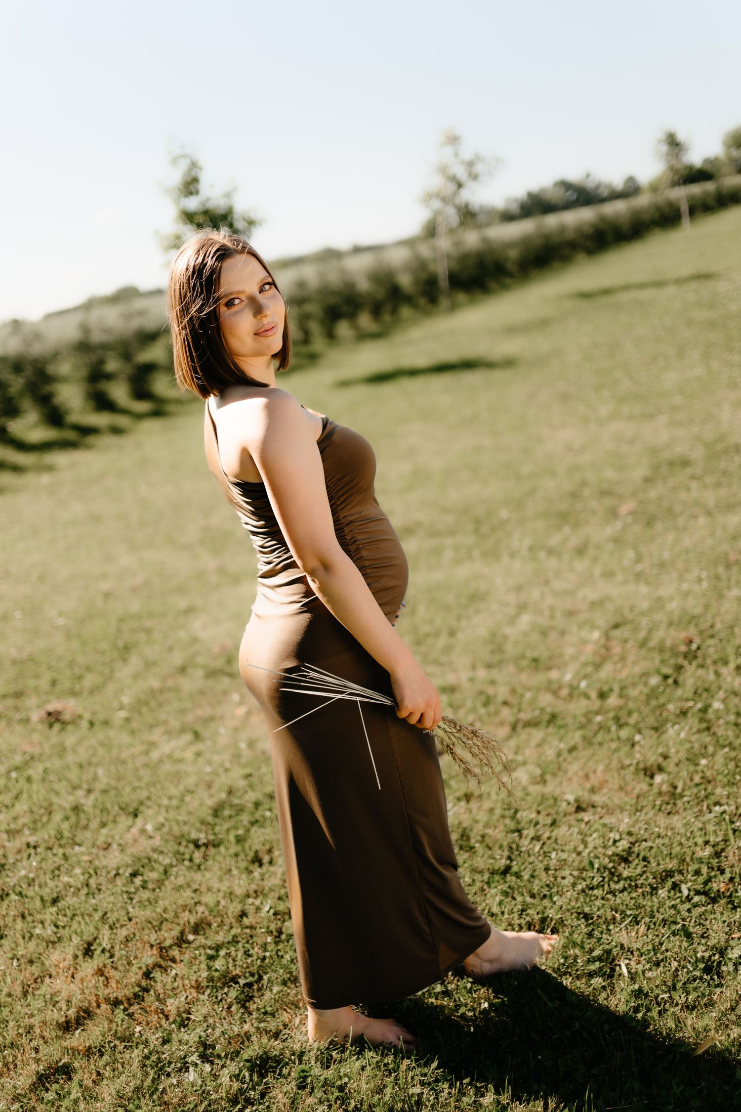Pregnant woman in brown maxi dress standing barefoot on green hillside in golden hour sunlight.