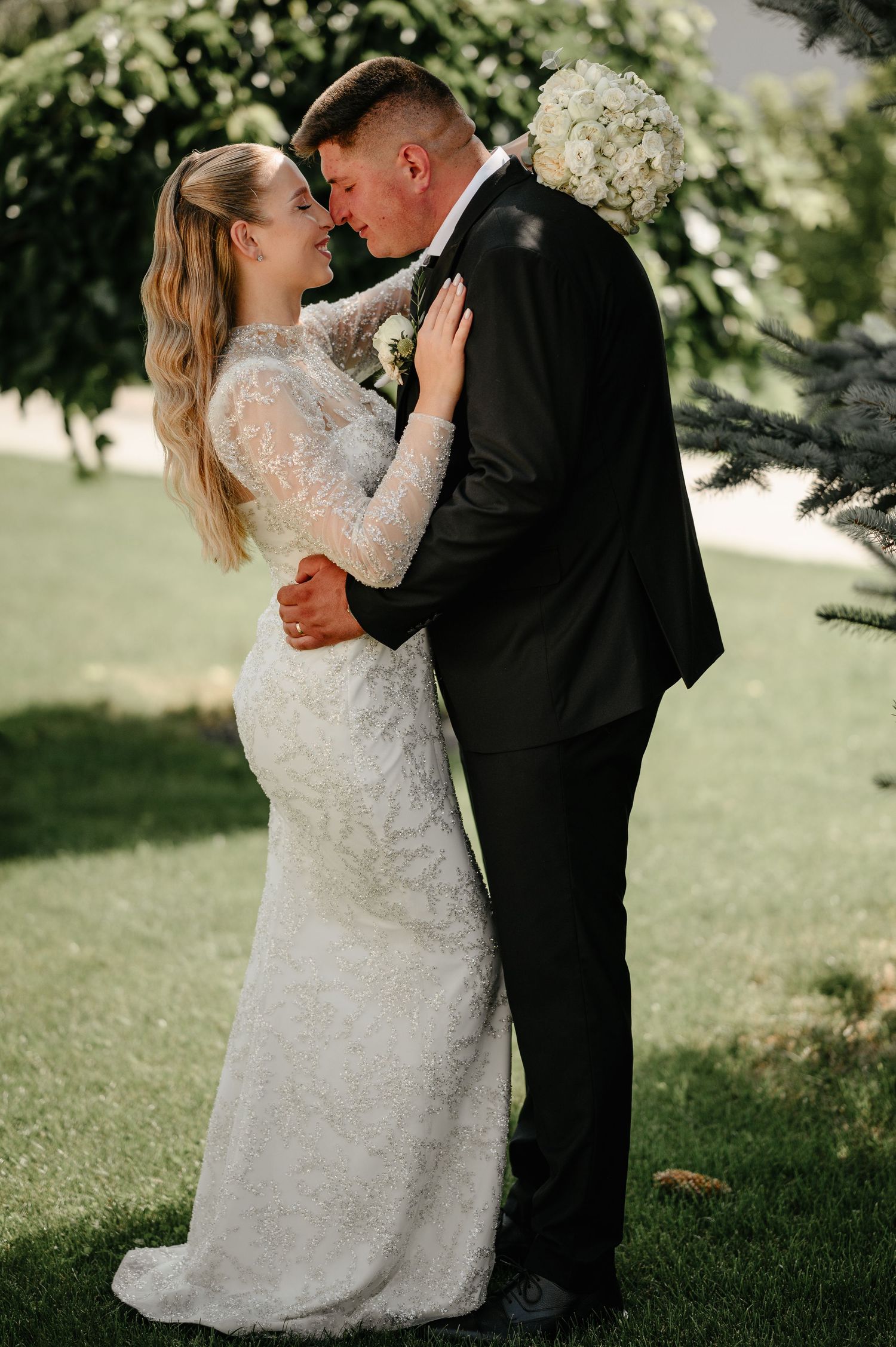 Bride in fitted lace gown and groom in black tuxedo sharing romantic moment on green lawn.