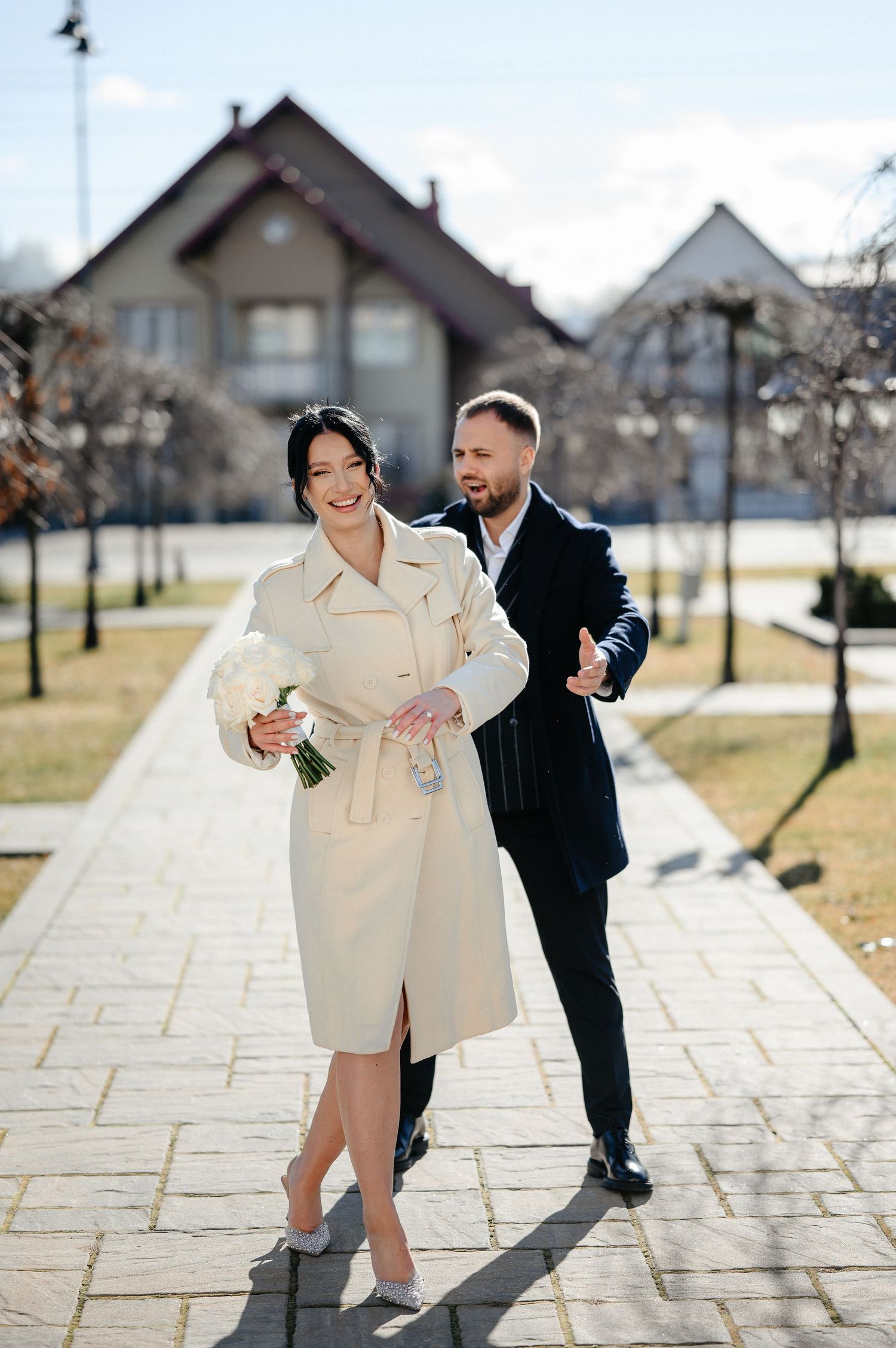Smiling bride in beige trench coat holding white bouquet walks with groom in dark suit on sunny outdoor path.