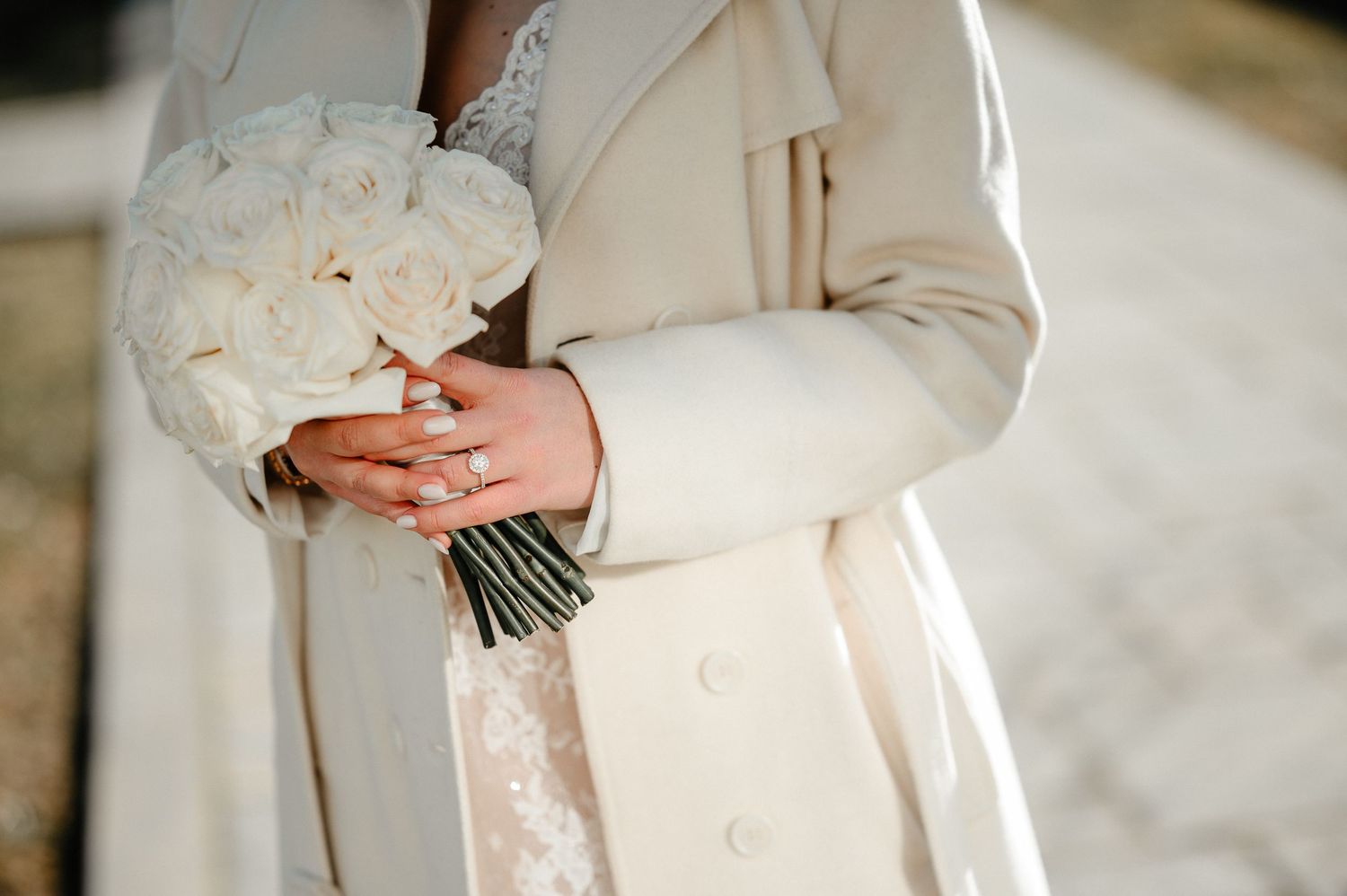 Bride holding white rose bouquet wearing diamond ring and elegant cream coat over lace wedding dress.