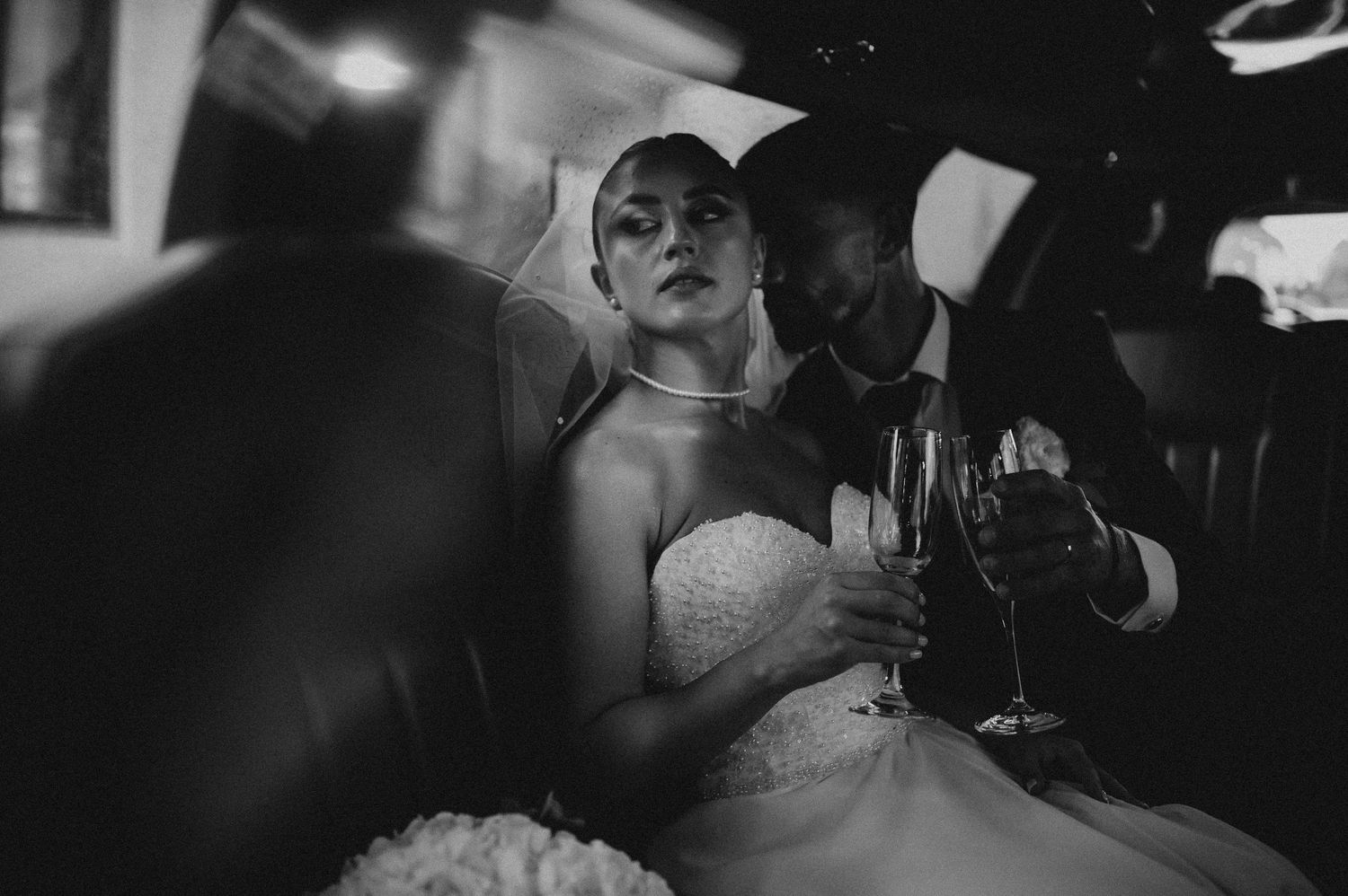 Black and white photo of a bride and groom sitting in a limousine, holding champagne glasses on their wedding day.