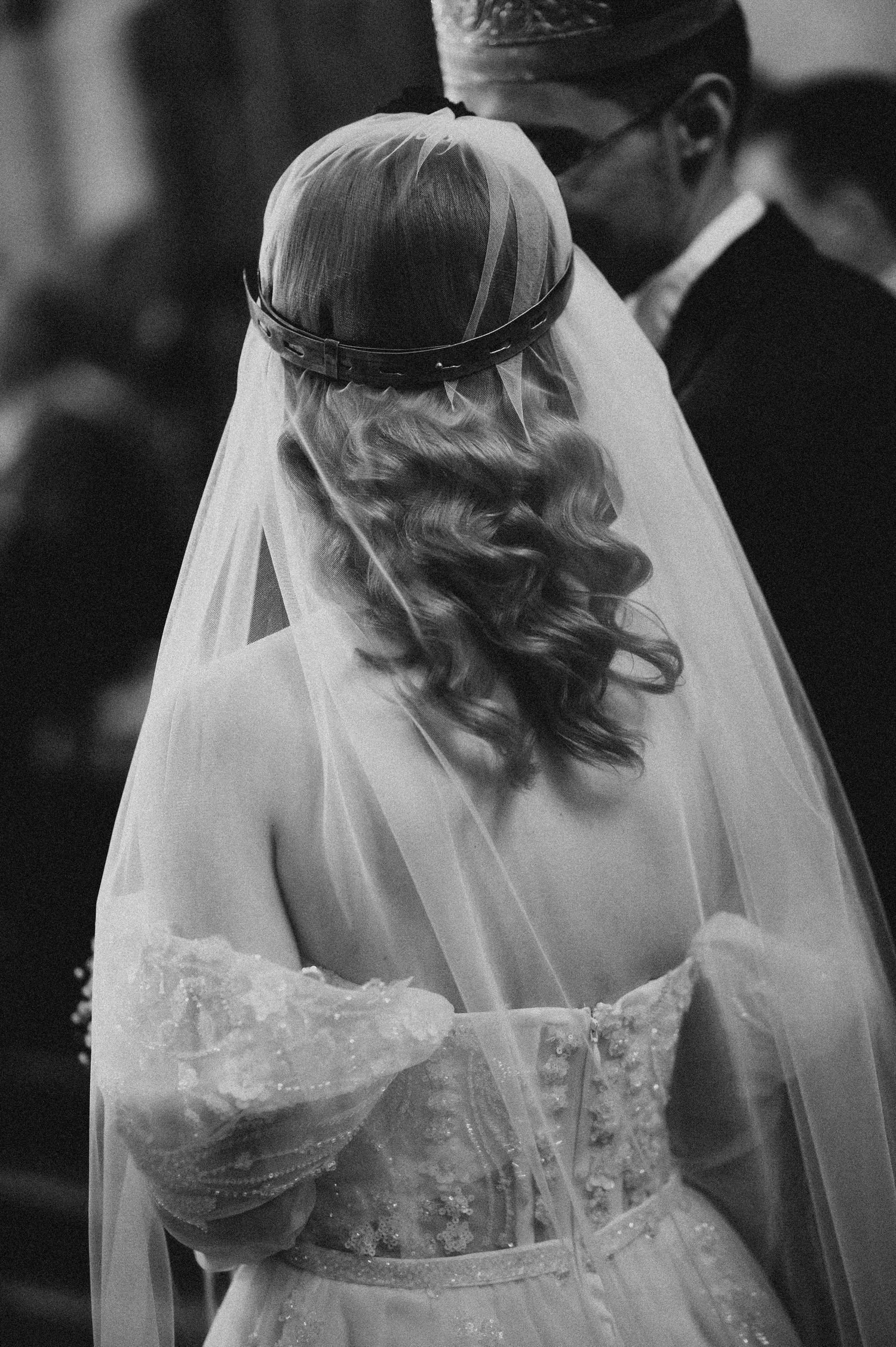 Black and white photo of a bride from behind, showing curly hair, floral crown, and elegant lace wedding dress with veil.