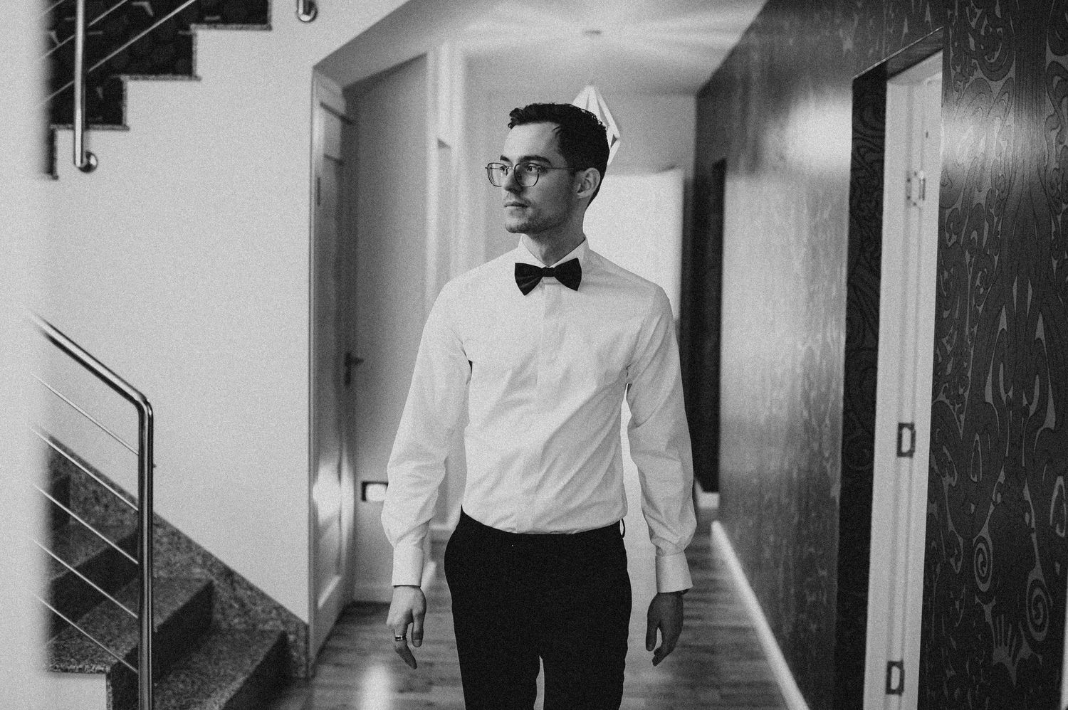 Elegant groom in white dress shirt and black bow tie standing in a modern hallway, black and white photo.