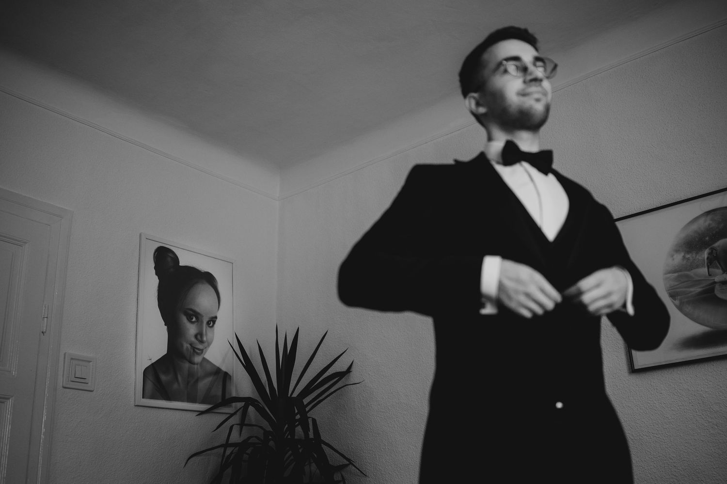 Man in tuxedo adjusting jacket in black and white photo with portrait painting on wall behind him.