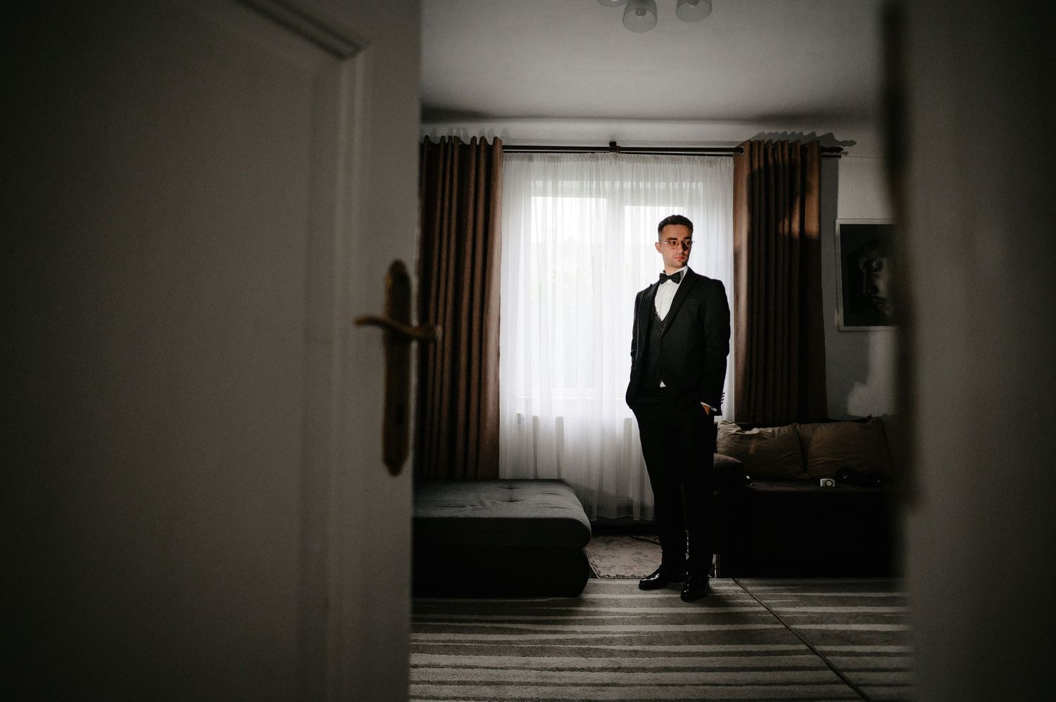 Groom in black tuxedo standing in hotel room by window before wedding ceremony.